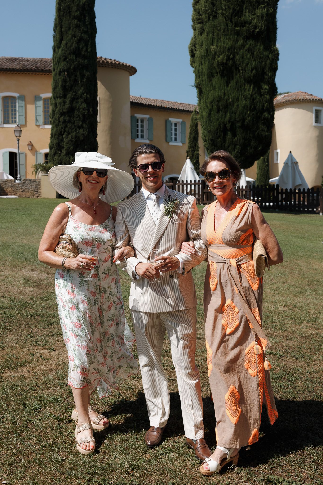 A groom in a cream suit poses with two elegantly dressed women at an outdoor wedding venue in the south of France.