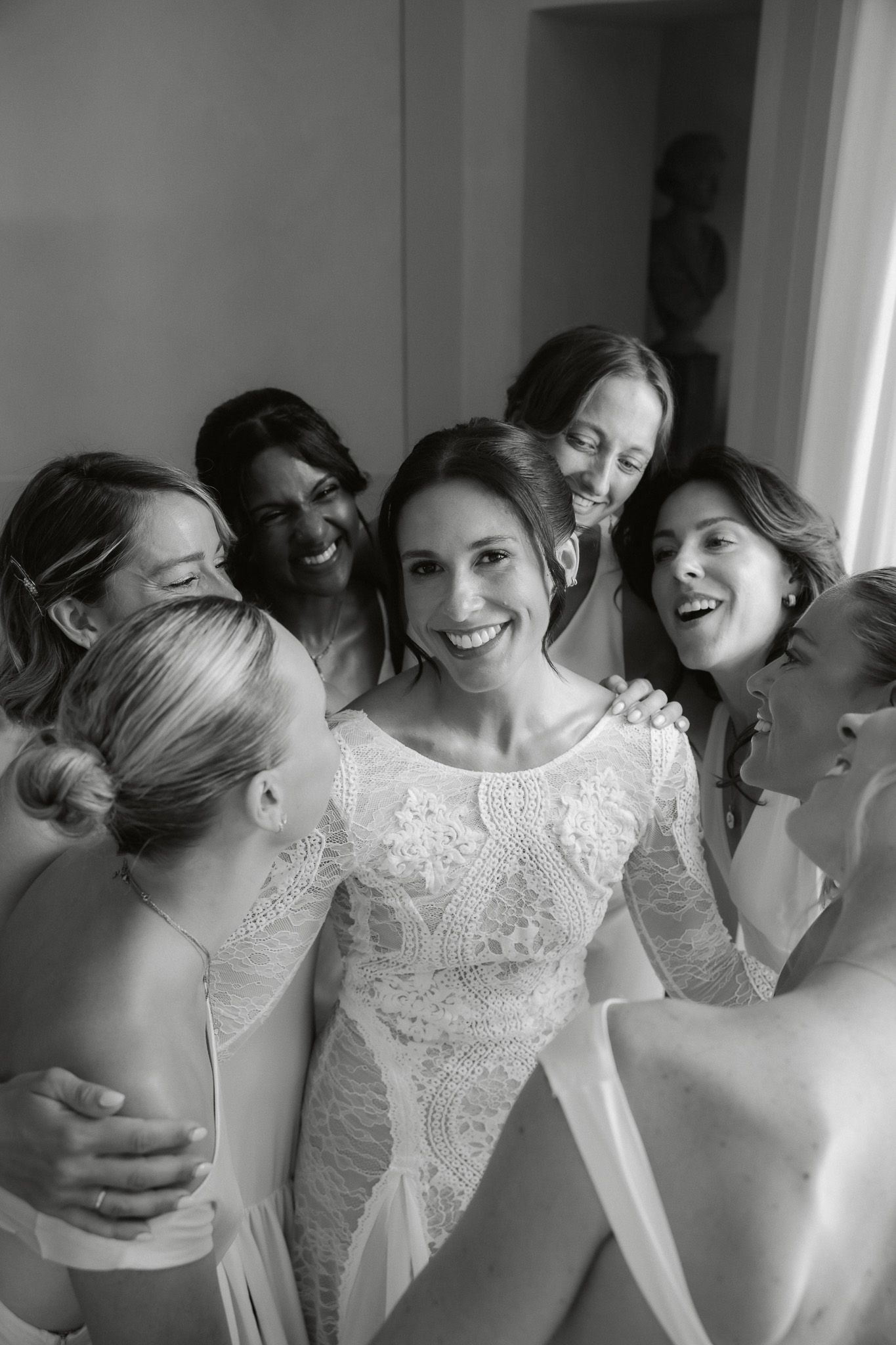 Black and white photo of a smiling bride in lace gown surrounded by her bridesmaids sharing a joyful moment.