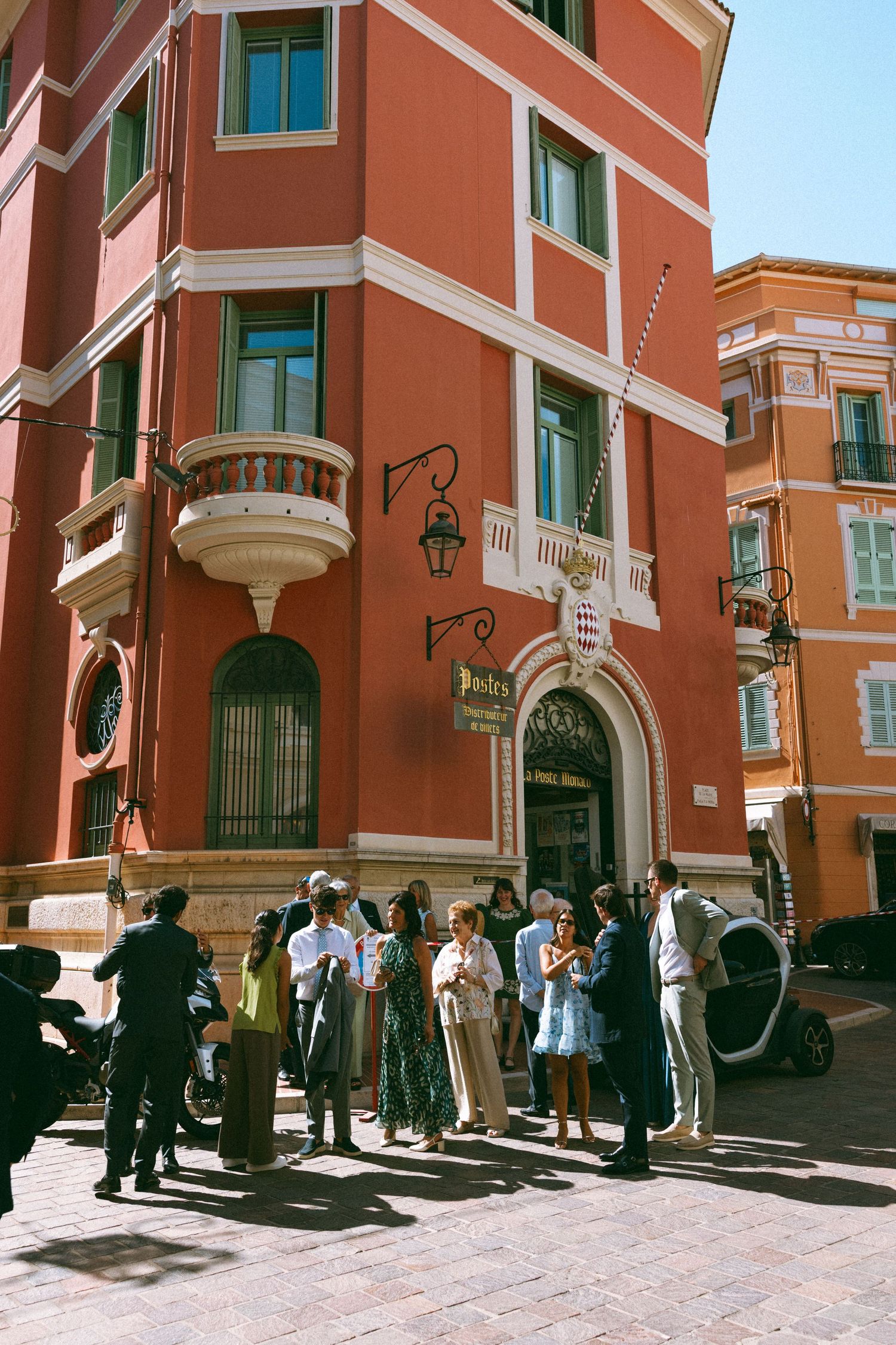 Group of people gathered outside a vibrant red building on a sunny cobblestone street in Europe.