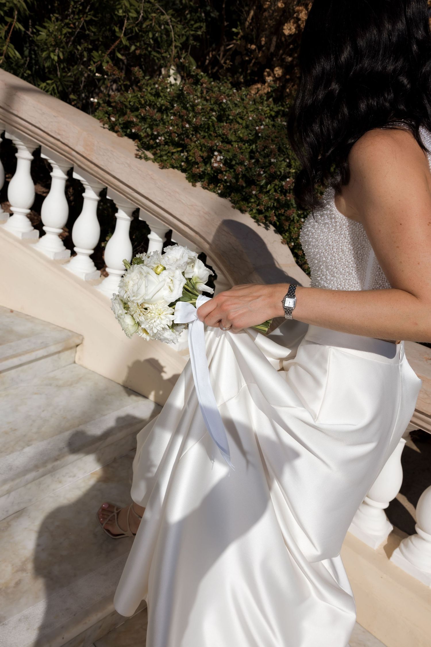 Bride in white satin gown holding bouquet of white roses while ascending elegant stone staircase.