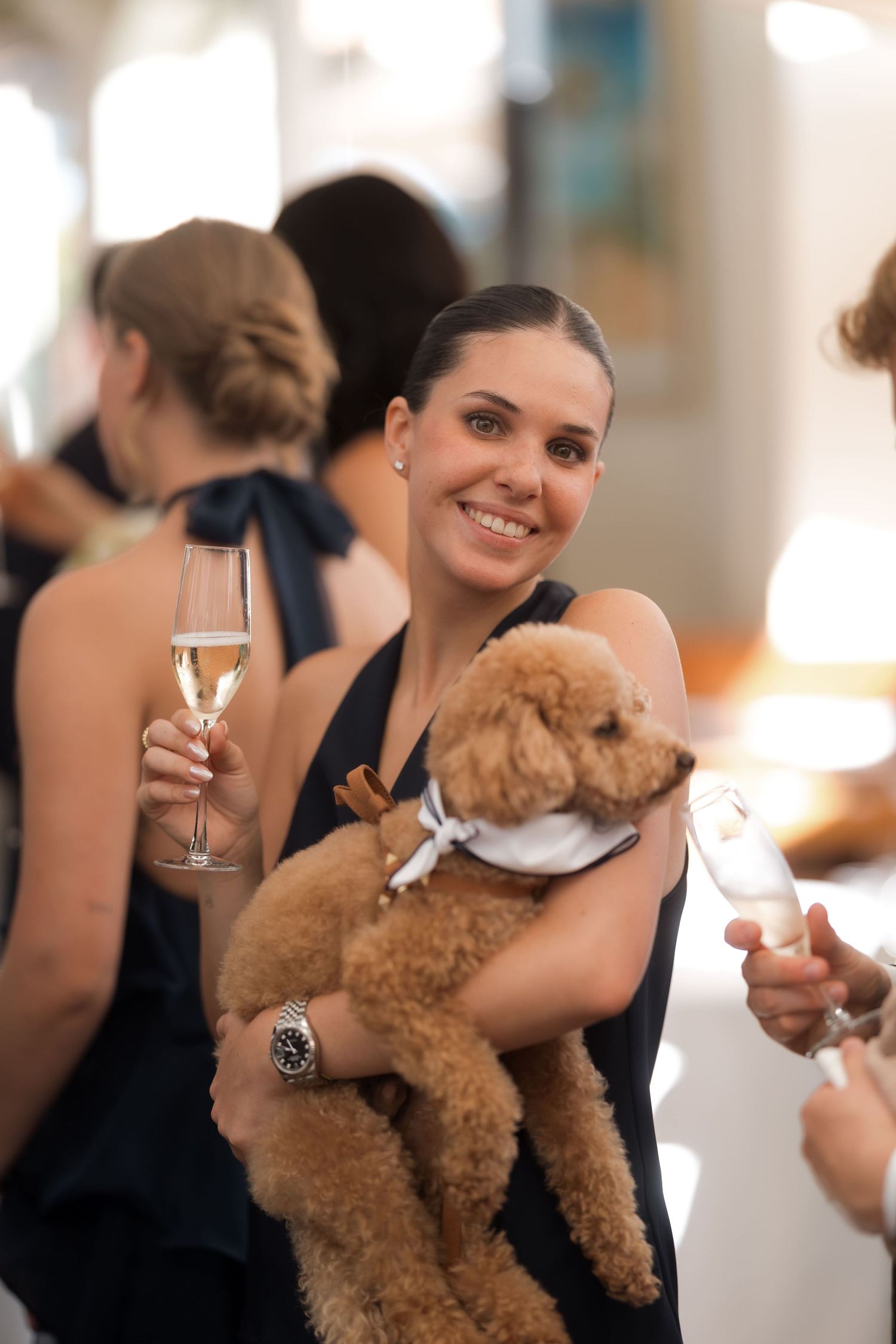 Smiling woman in black dress holding a fluffy dog and champagne glass at an elegant event.
