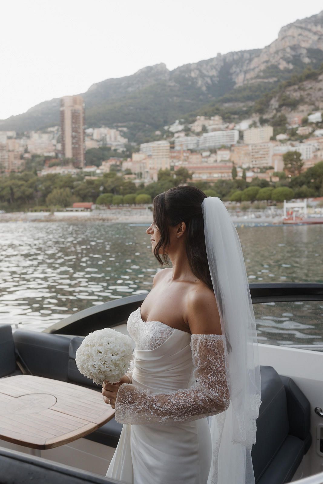 A bride in a white lace dress stands at the railing of a yacht overlooking Monaco's harbor and mountainous coastline.