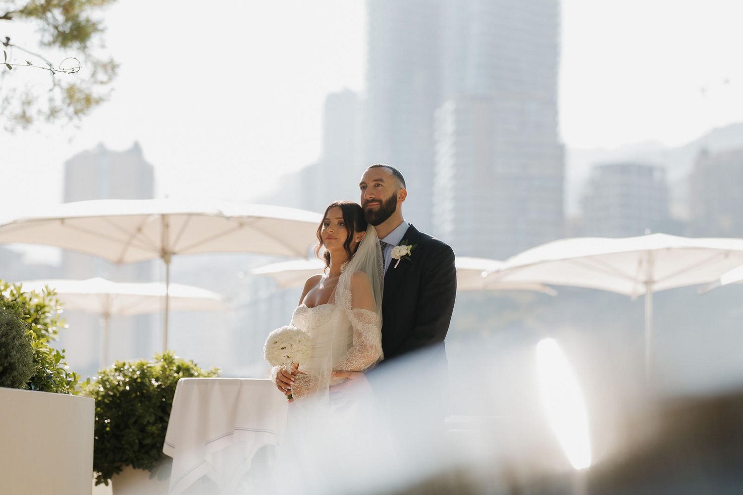 Wedding couple embraces under white umbrellas on a sunny day with city skyline in background.