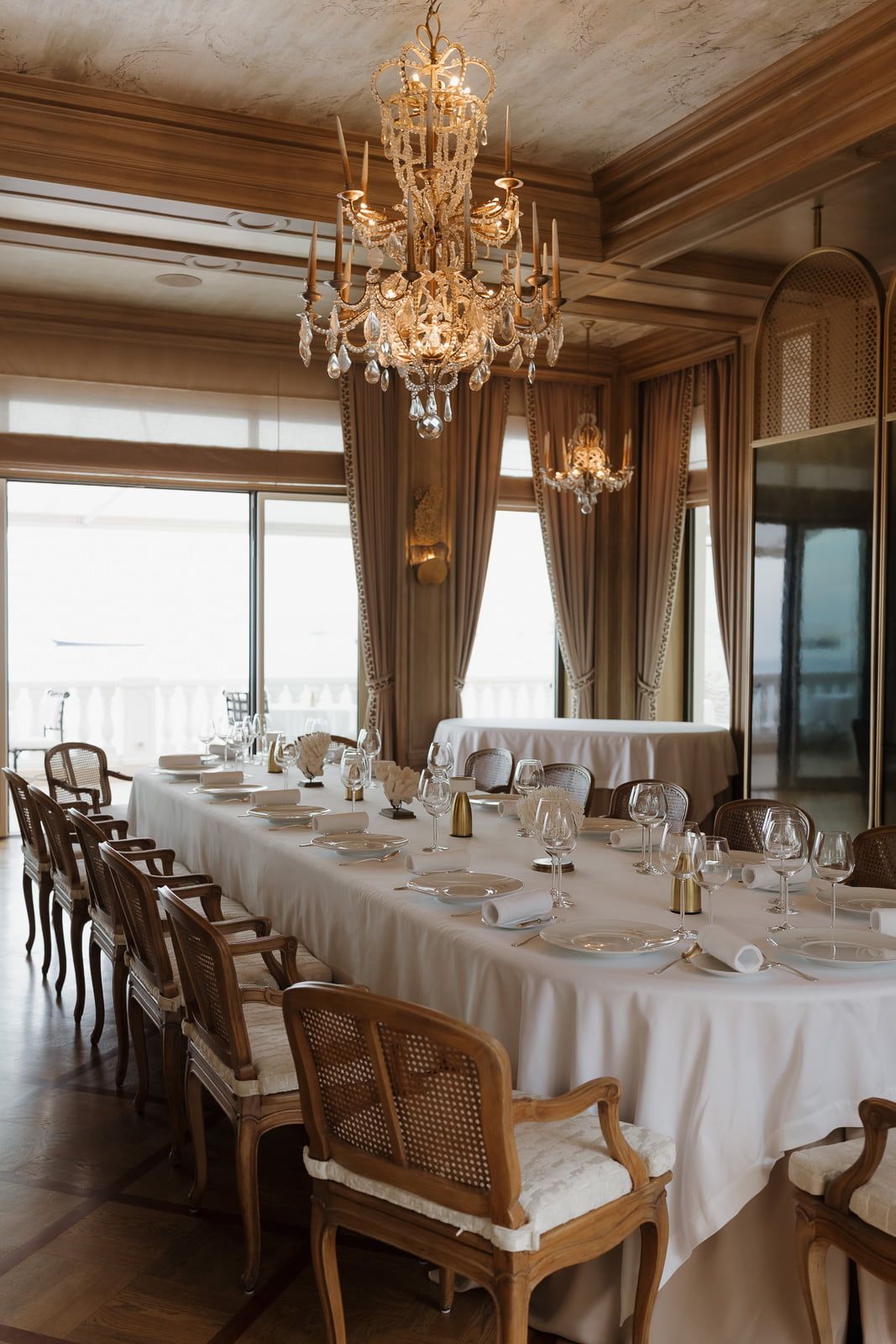 Elegant dining room with crystal chandeliers, white tablecloths, and cane-back chairs in a luxurious restaurant setting.