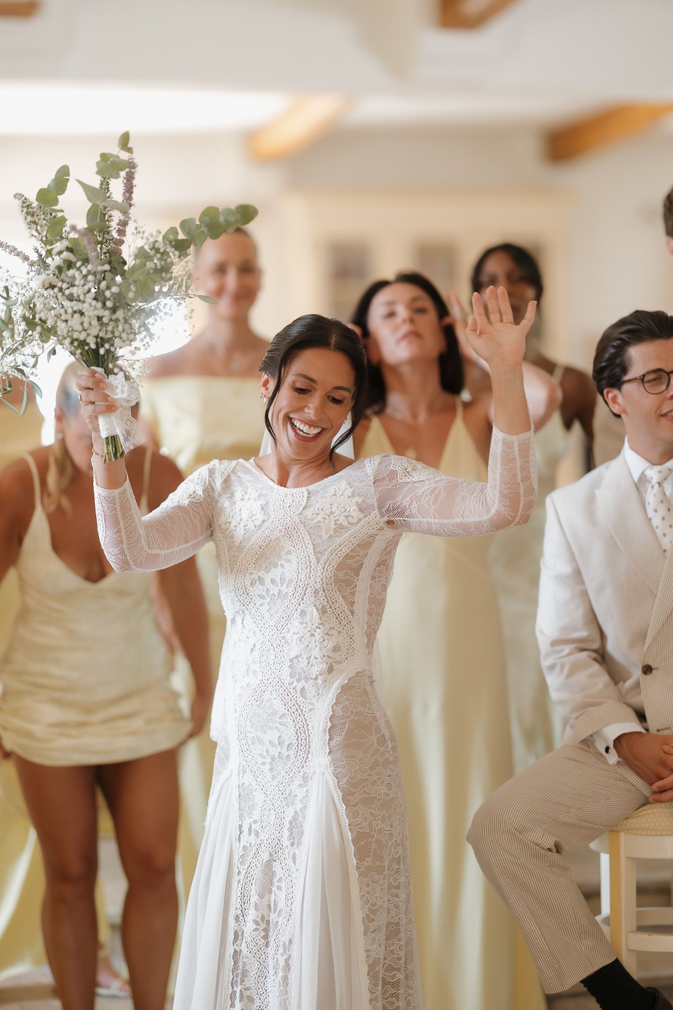 Smiling bride in lace gown holds bouquet aloft walking down aisle with bridesmaids in yellow dresses.