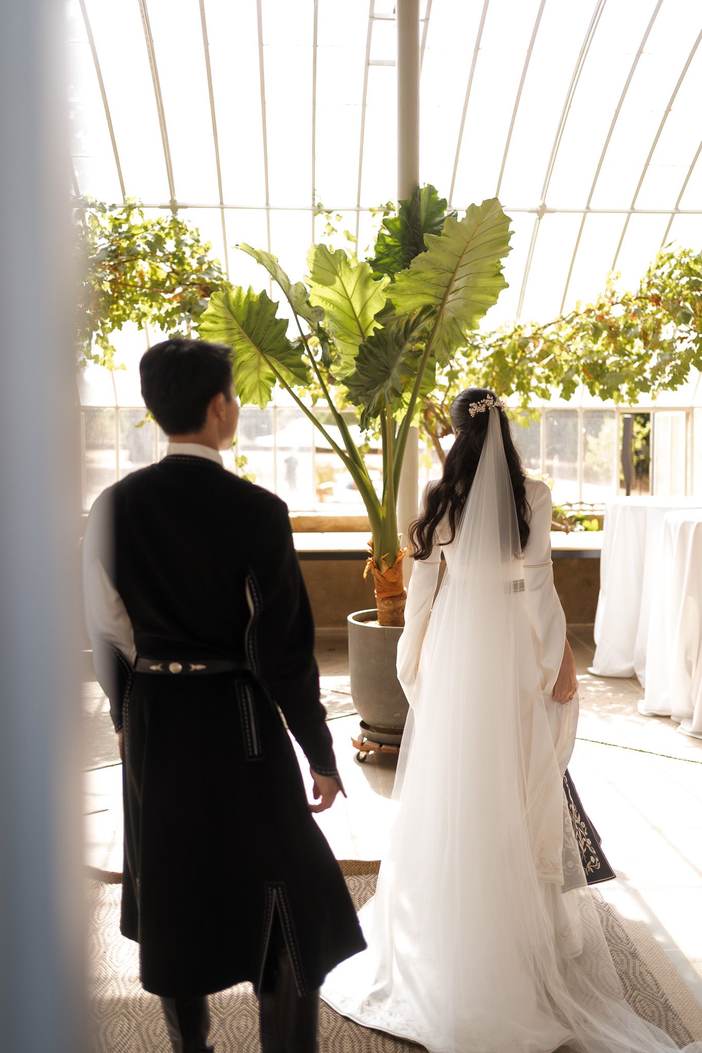 Bride in white gown and groom in black traditional attire stand together in sunlit greenhouse venue.