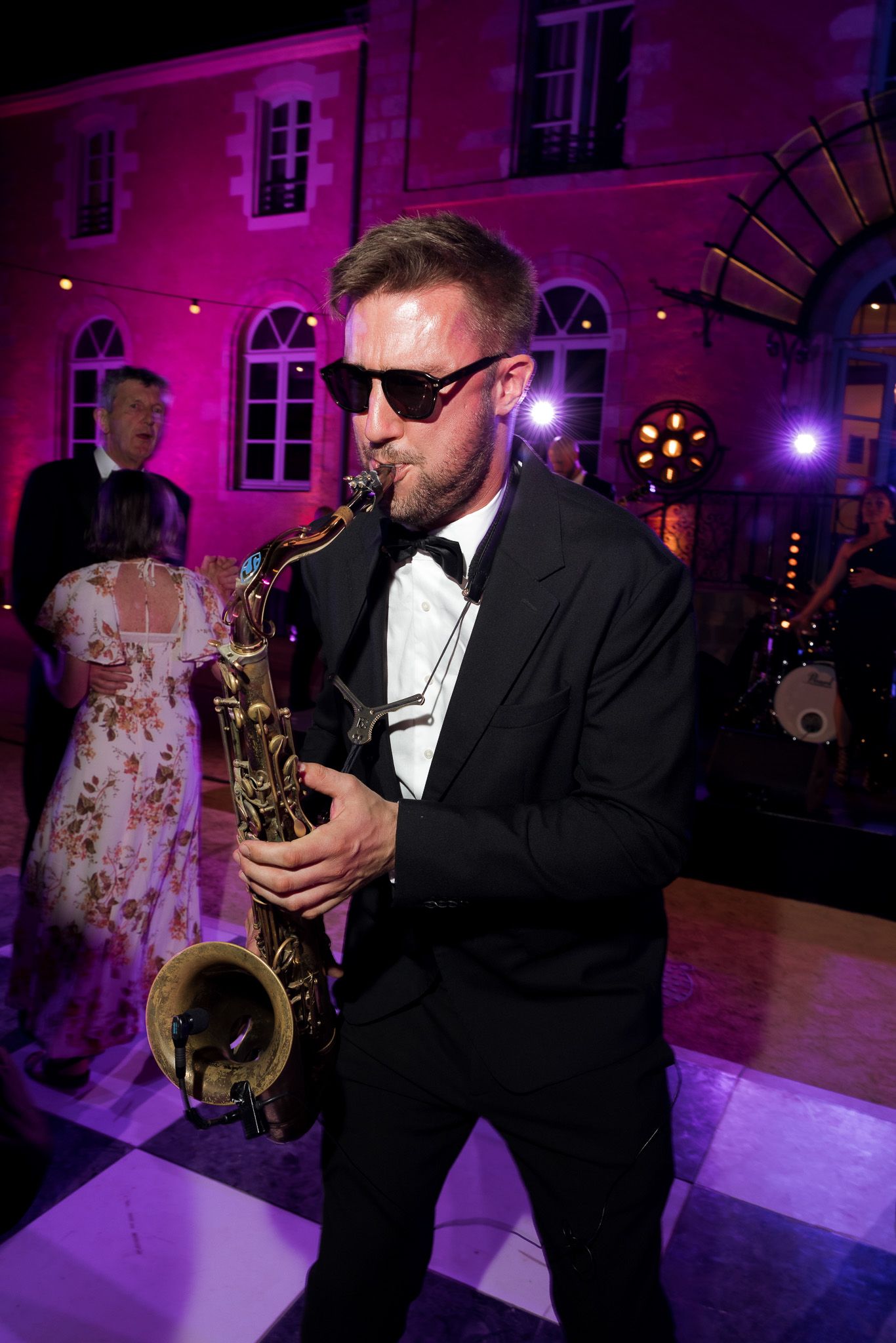 A saxophonist in a tuxedo and sunglasses performs at an elegant outdoor evening event with pink lighting.
