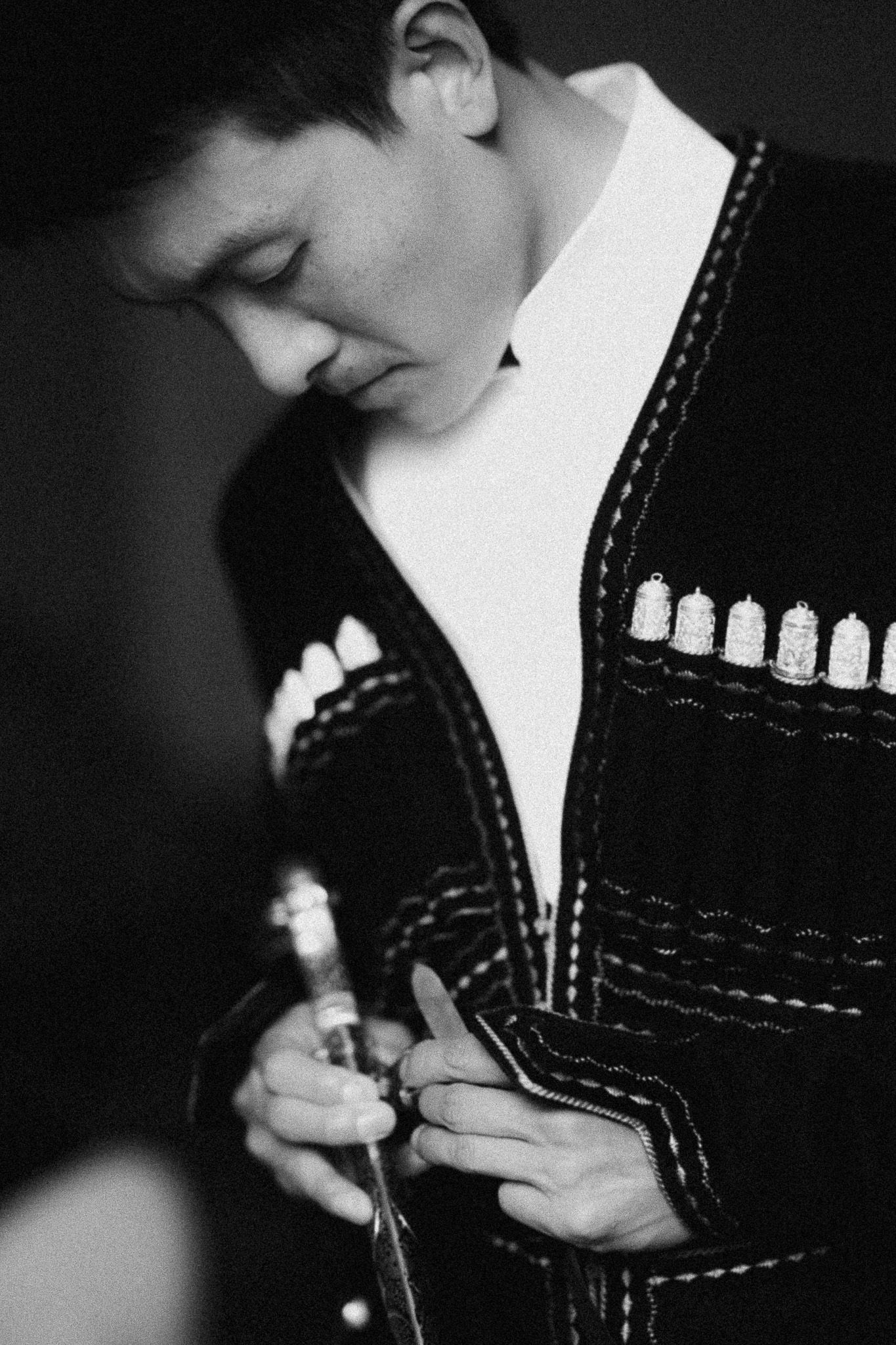 Black and white photo of a young man in embroidered jacket looking down at his hands holding a small object.