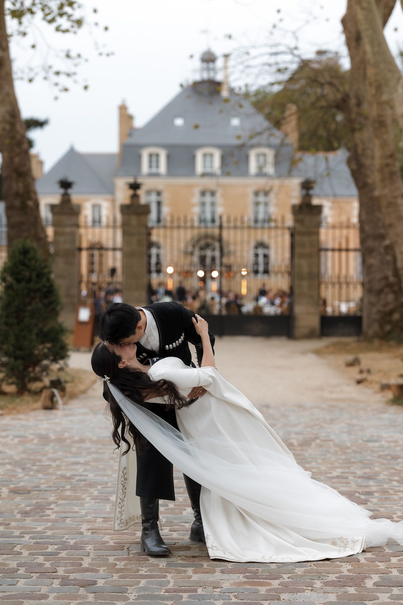 A couple shares a romantic dip kiss on a cobblestone path before an elegant French château at their wedding.