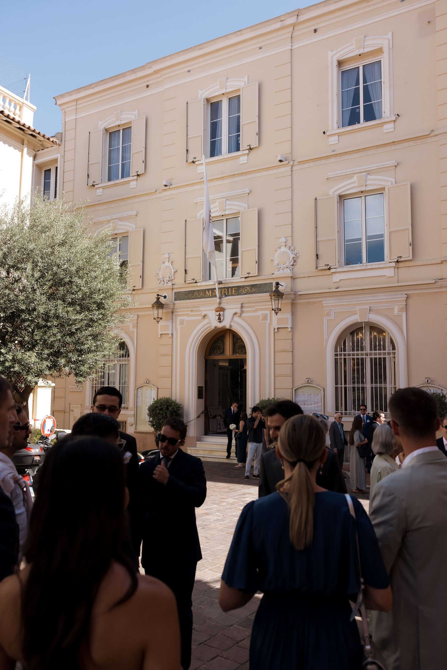 Elegant guests gather outside a classic French building with shuttered windows on a sunny day.
