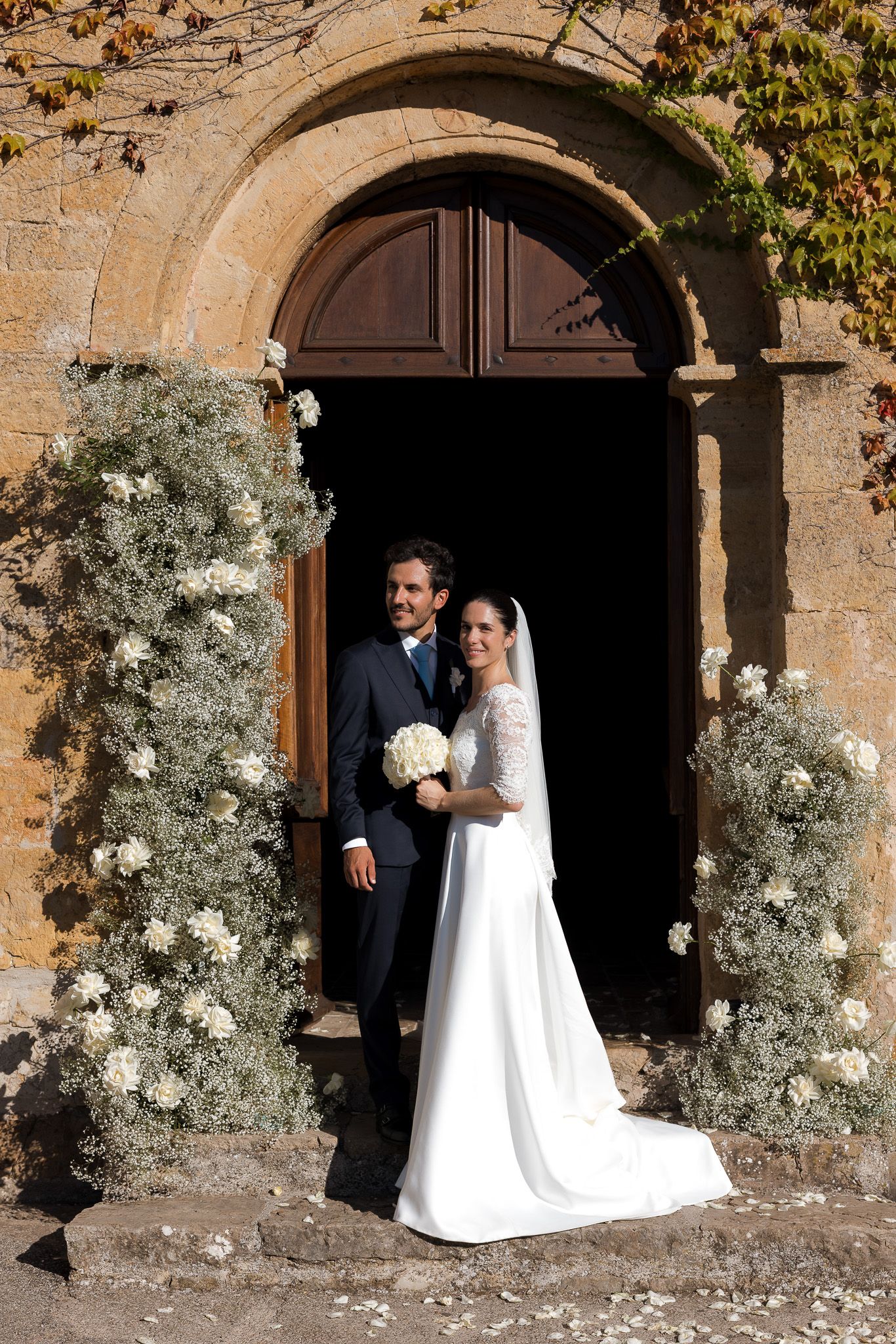 Bride and groom posing in a floral-adorned church doorway, bride in white gown holding bouquet, groom in navy suit.