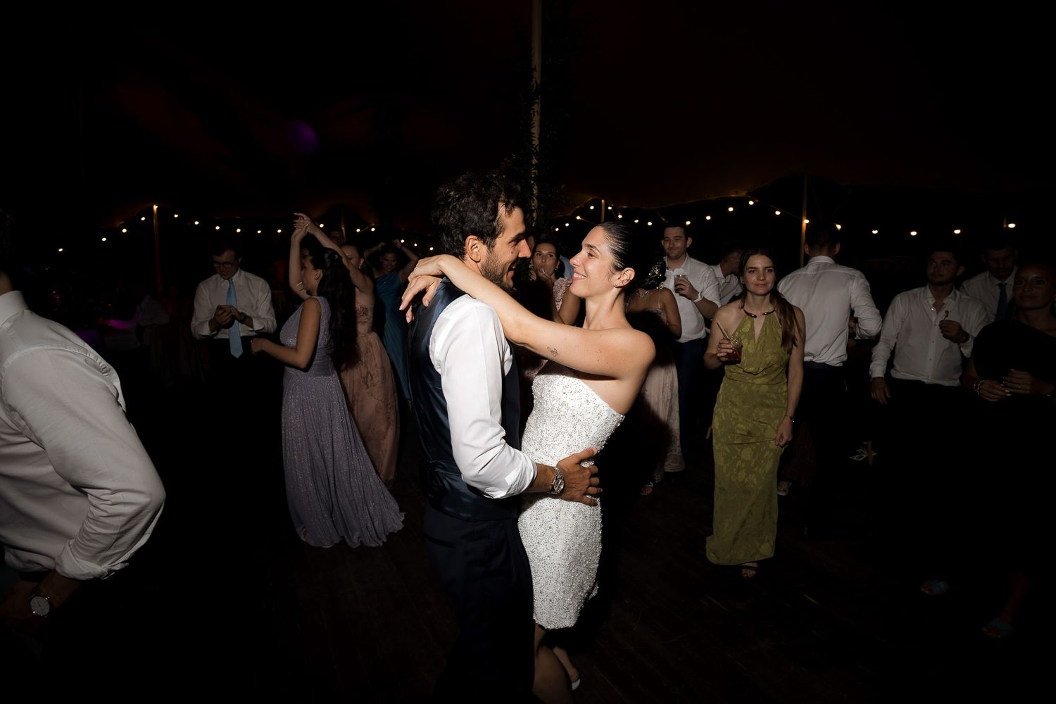 Bride and groom share romantic first dance surrounded by guests under warm string lights at wedding reception.