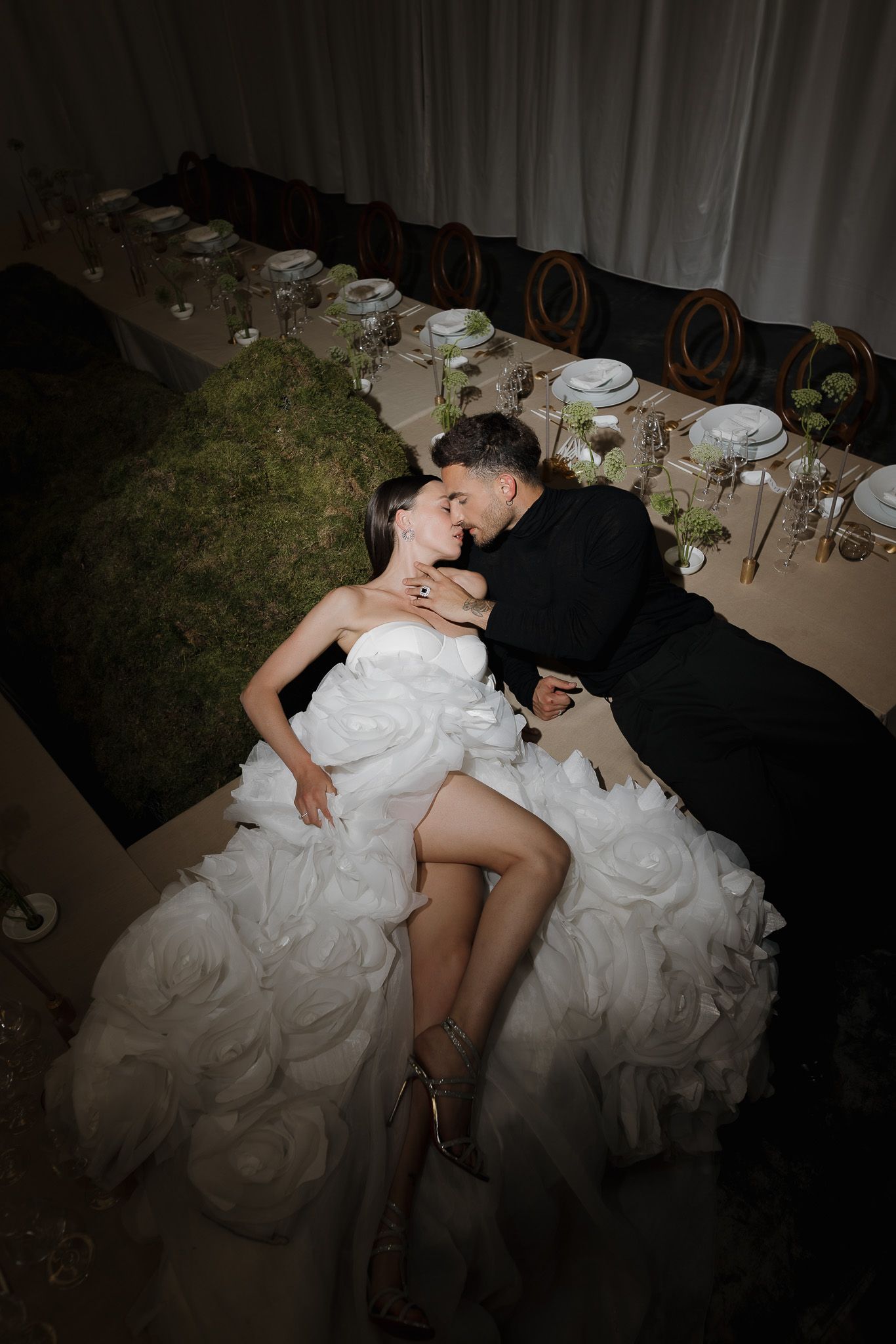 Bride in white ruffled gown and groom in black suit share romantic kiss while reclining beside elegantly set reception table.