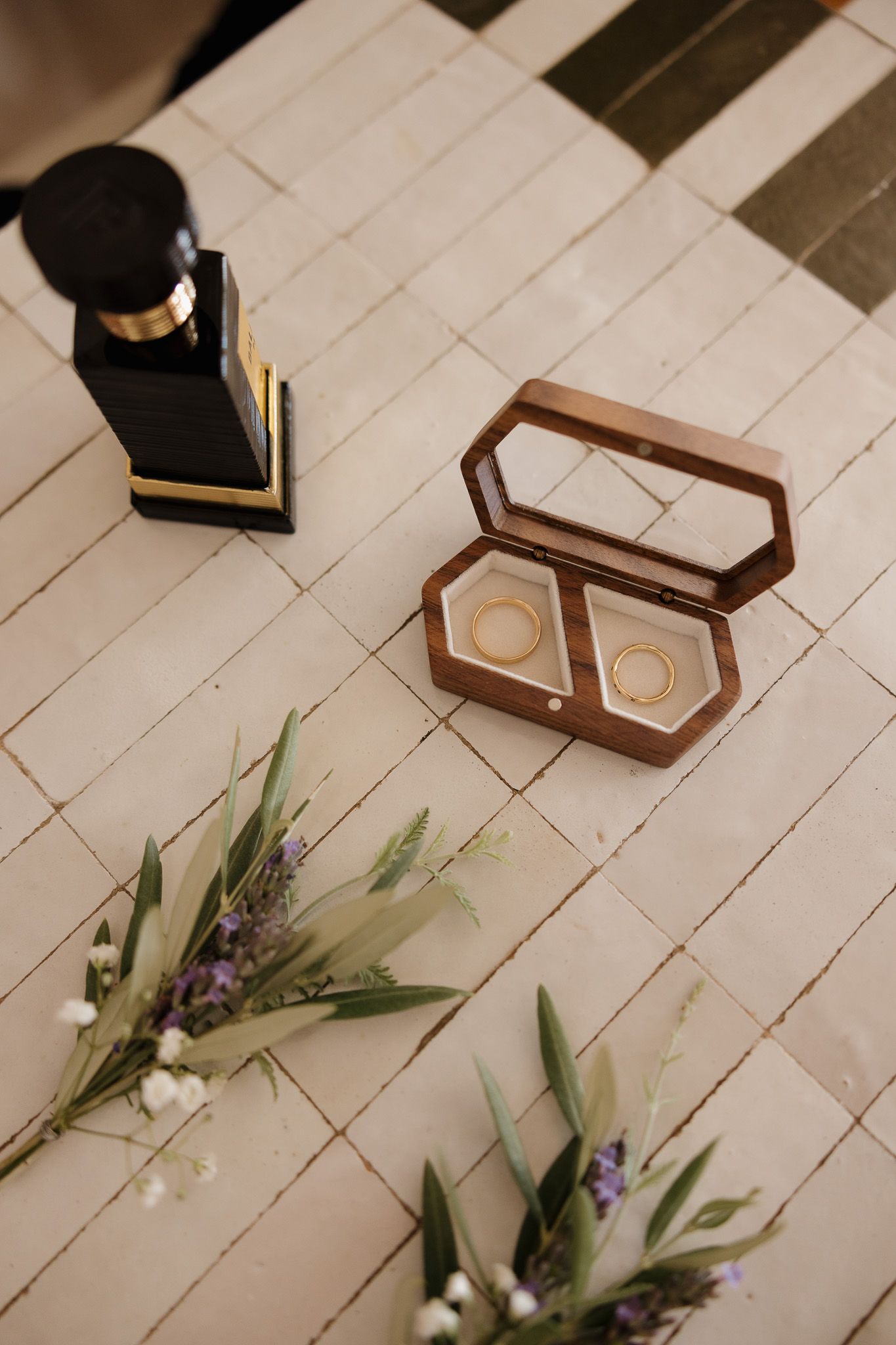 Two gold wedding rings in an open wooden ring box on beige tiles, with flowers and a perfume bottle nearby.