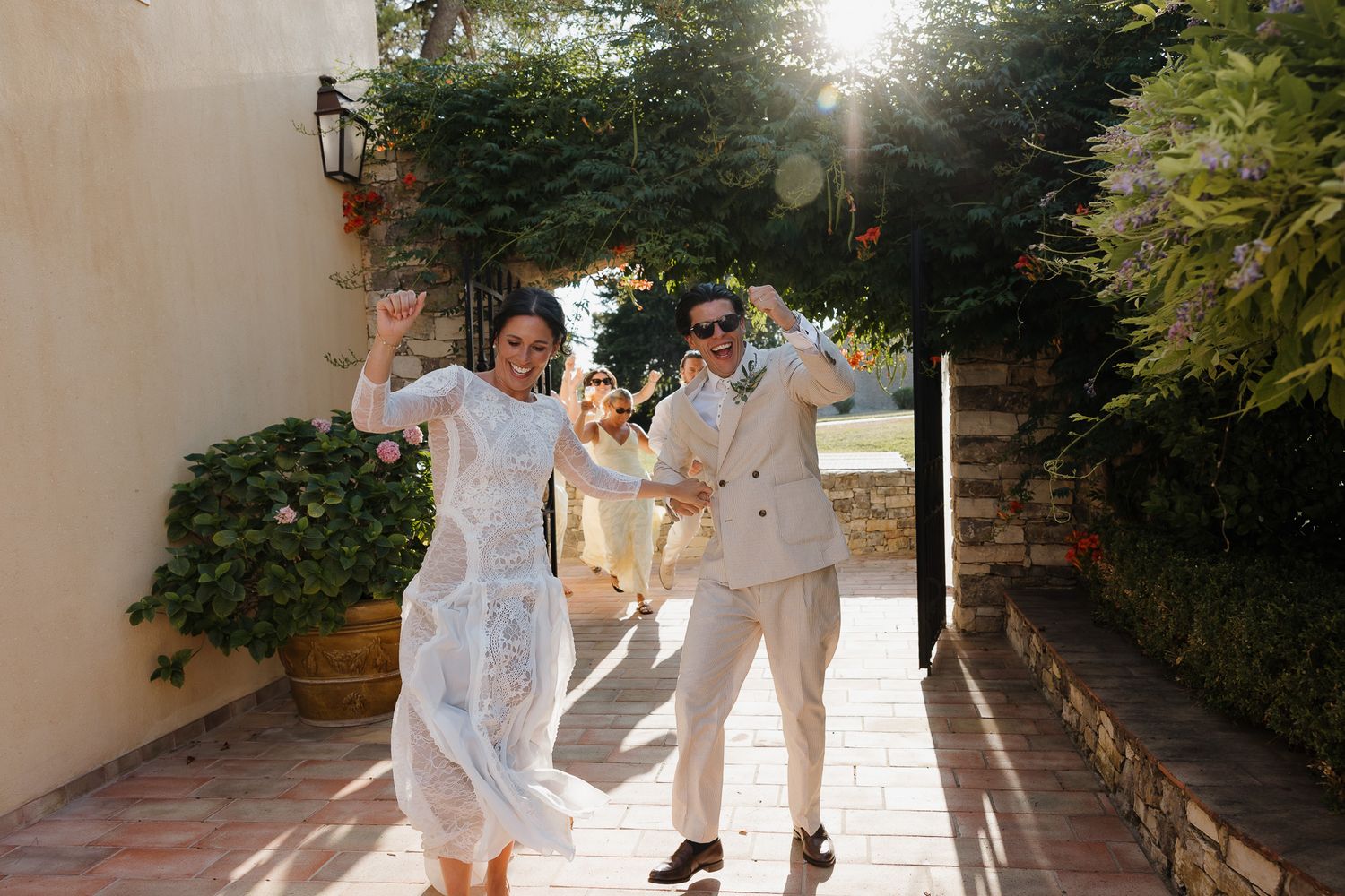 Joyful newlyweds in white outfits celebrate outdoors on a sunny patio surrounded by lush greenery.