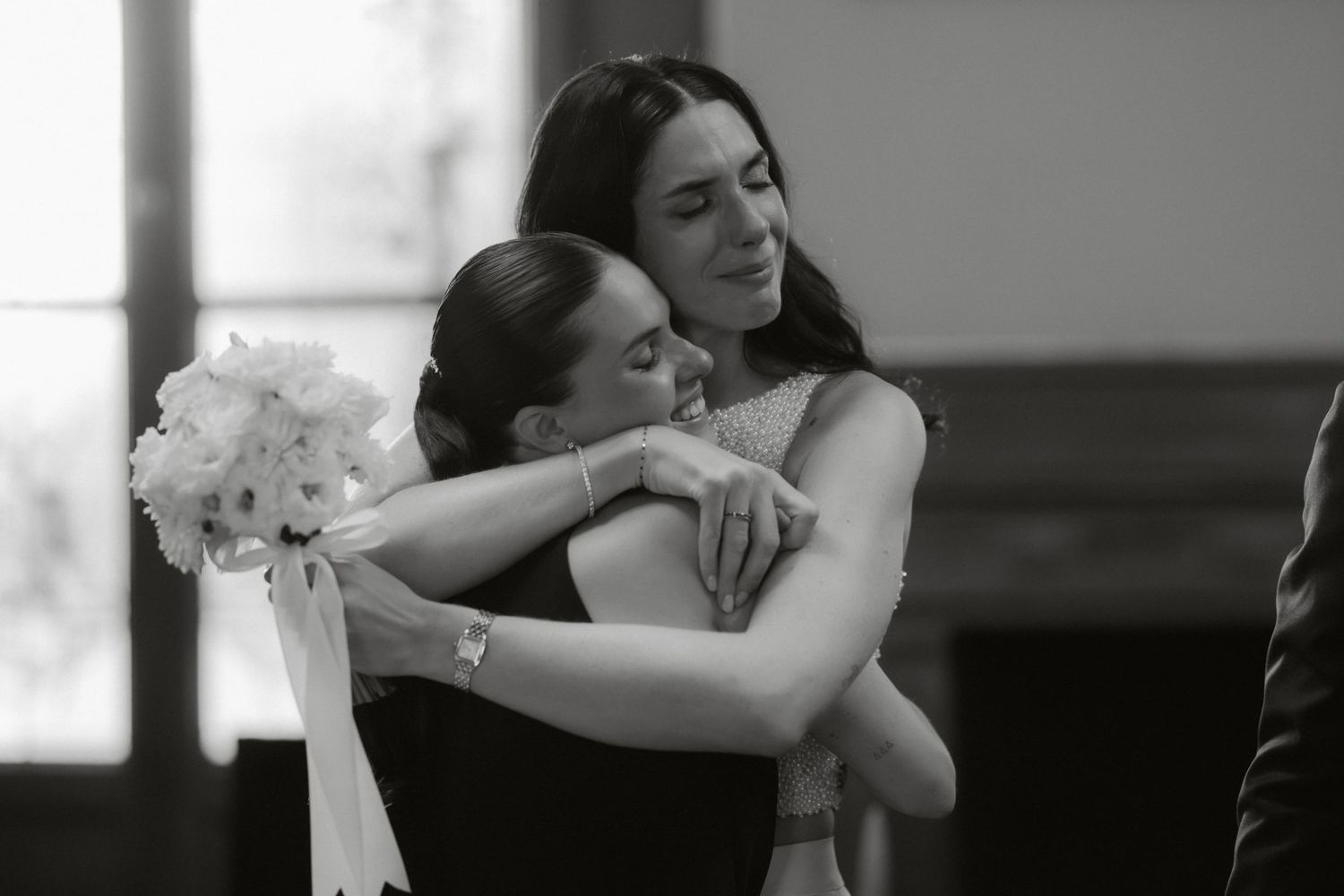 Two women share an emotional hug at a wedding, one holding a white bouquet, captured in black and white.