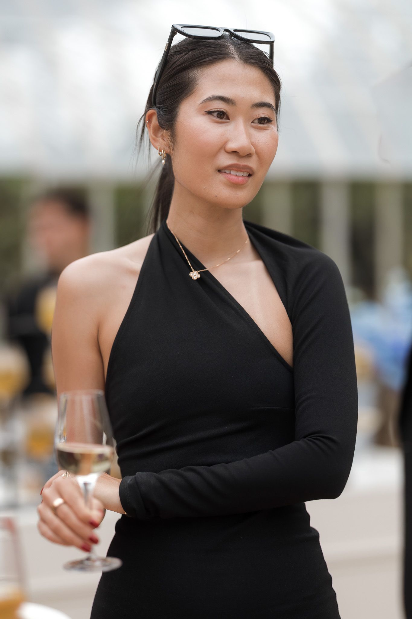 A young Asian woman in a black one-shoulder dress holds a champagne glass at an outdoor event.