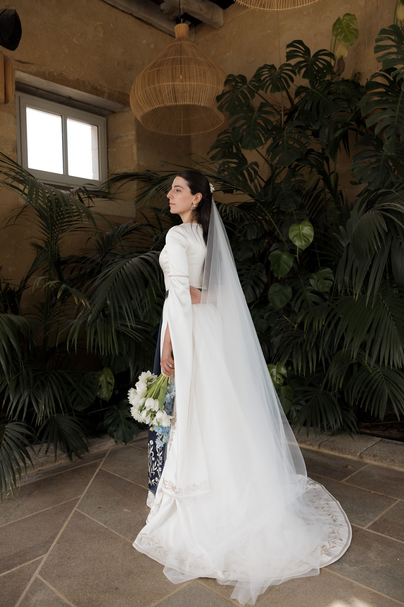 Bride in elegant white wedding gown with long veil holding bouquet, standing before lush tropical plants indoors.
