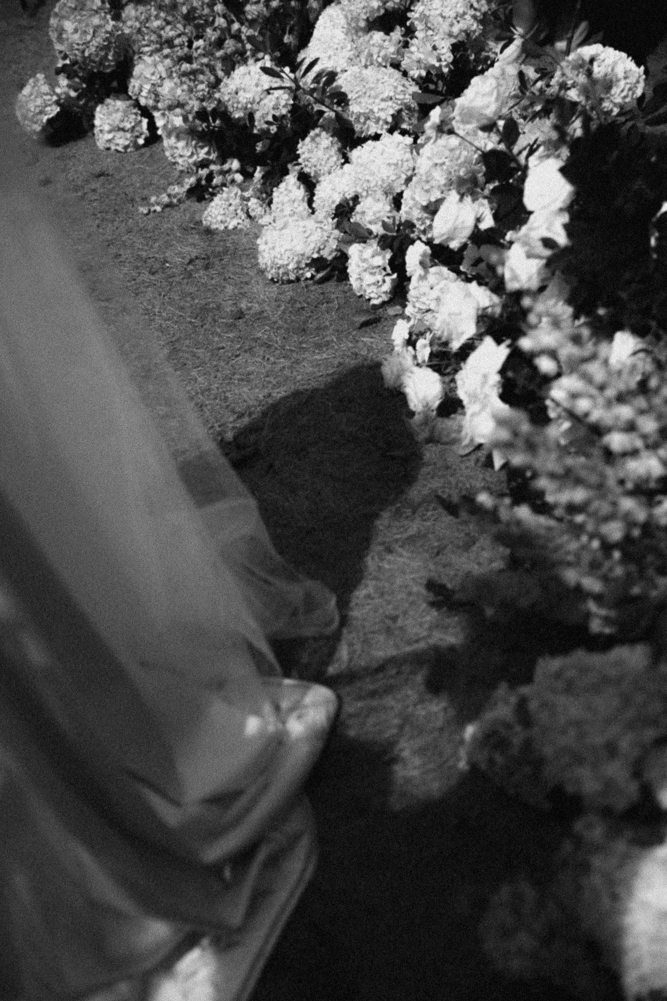 Black and white photo of a bride's gown and shoes beside lush white floral arrangements at a wedding ceremony.