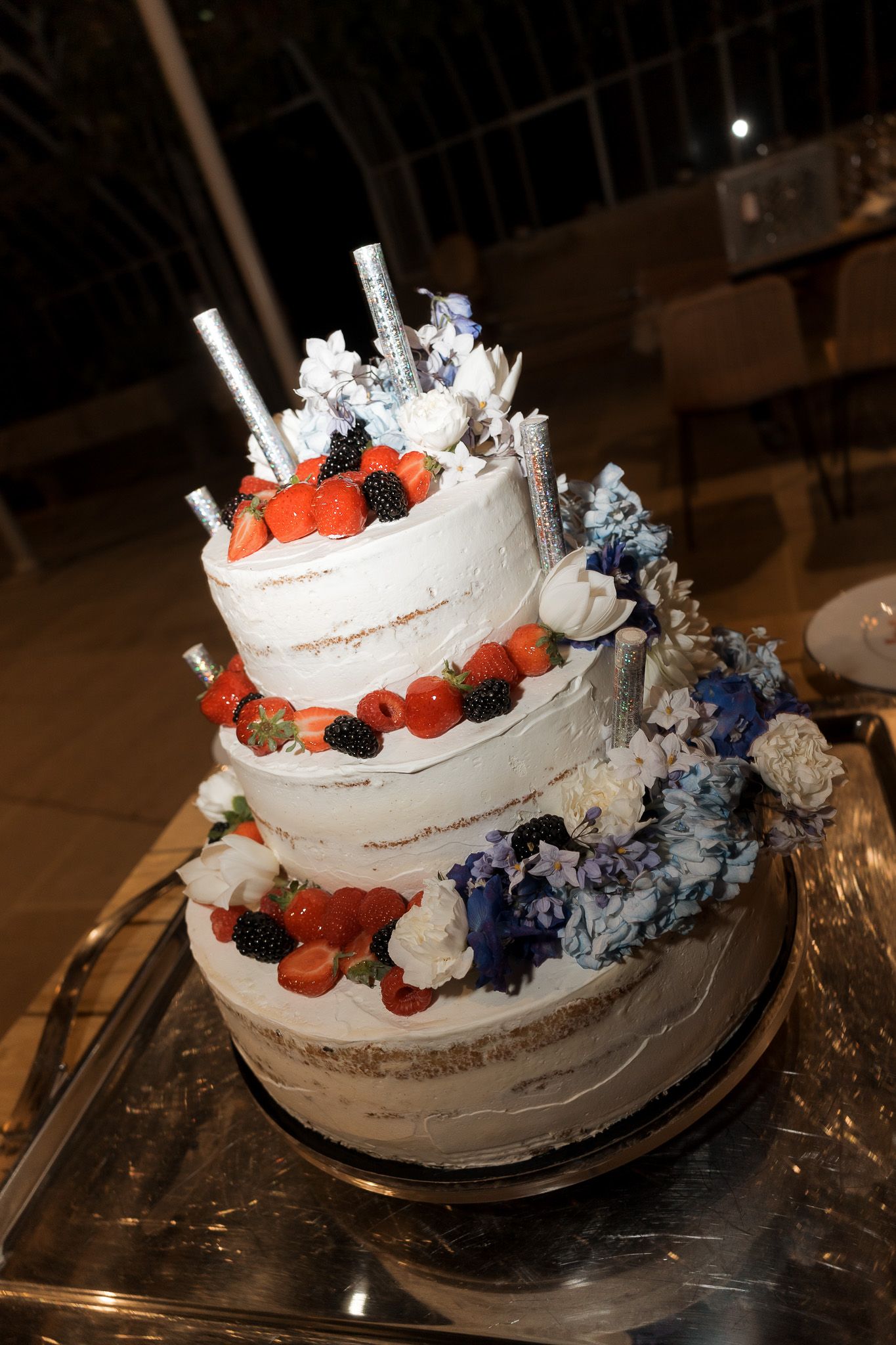 Three-tier naked wedding cake with fresh strawberries, blackberries, blue hydrangeas, and silver candles on a silver stand.