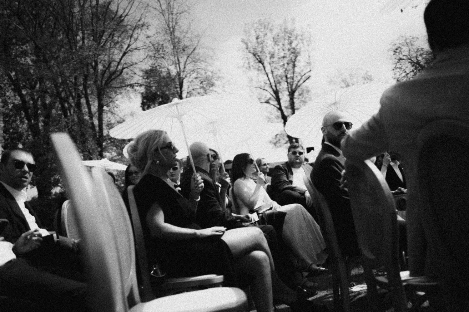 Black and white photo of elegantly dressed guests seated outdoors at a formal event, with trees visible in background.