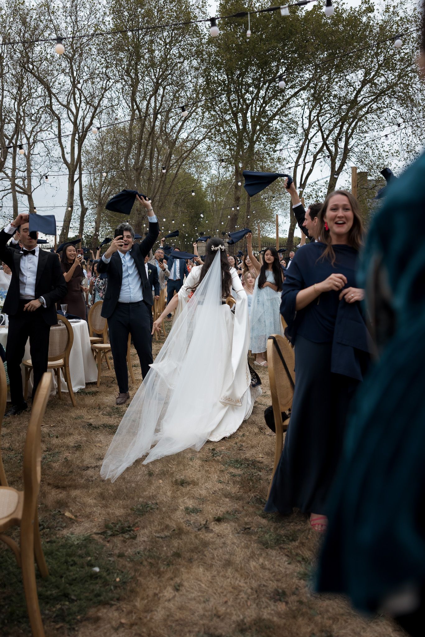 Bride in white gown walks down outdoor aisle as guests celebrate at a joyful wedding ceremony.