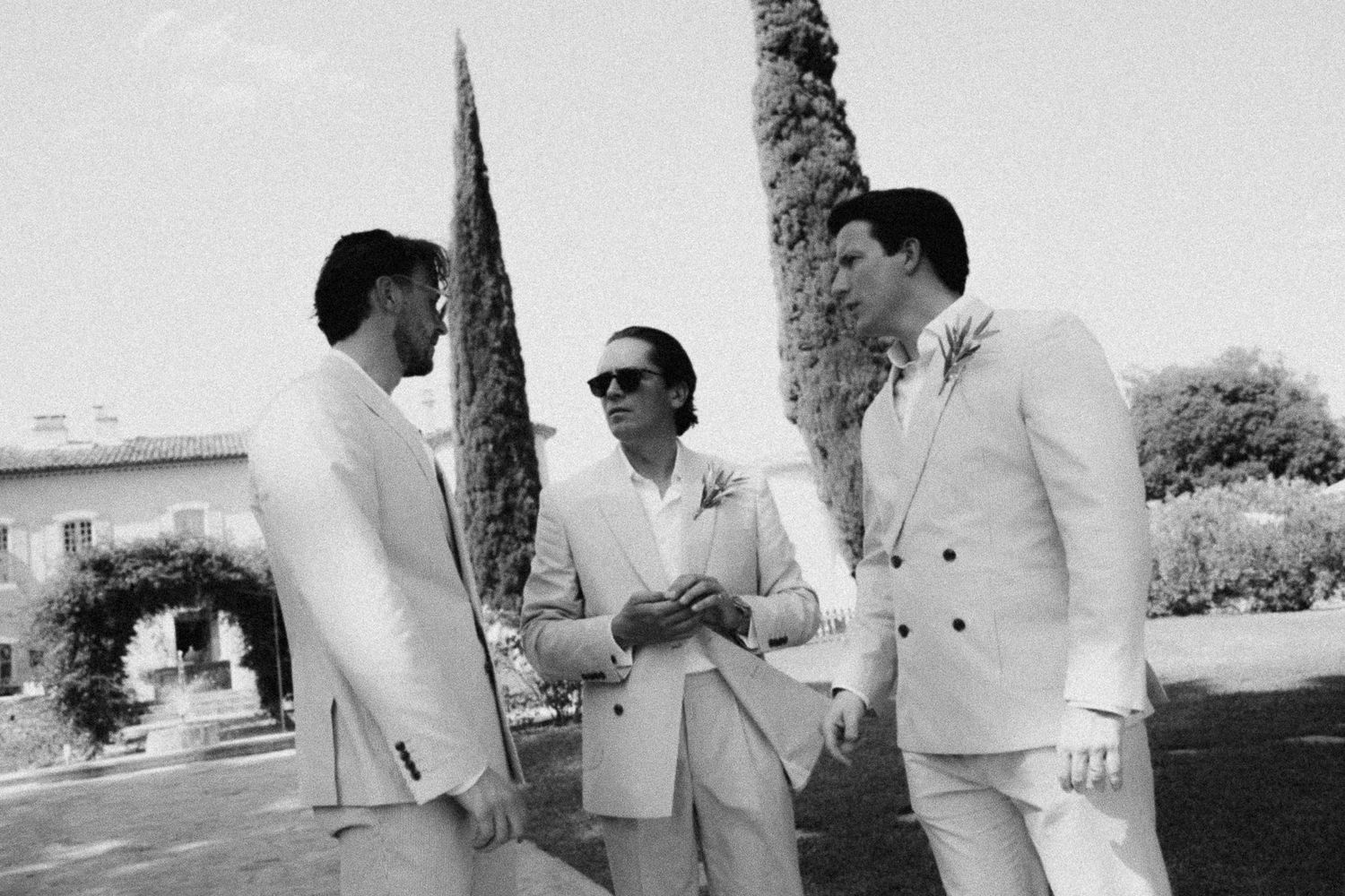 Three men in white suits socializing outdoors at a wedding venue with cypress trees and a stone building.