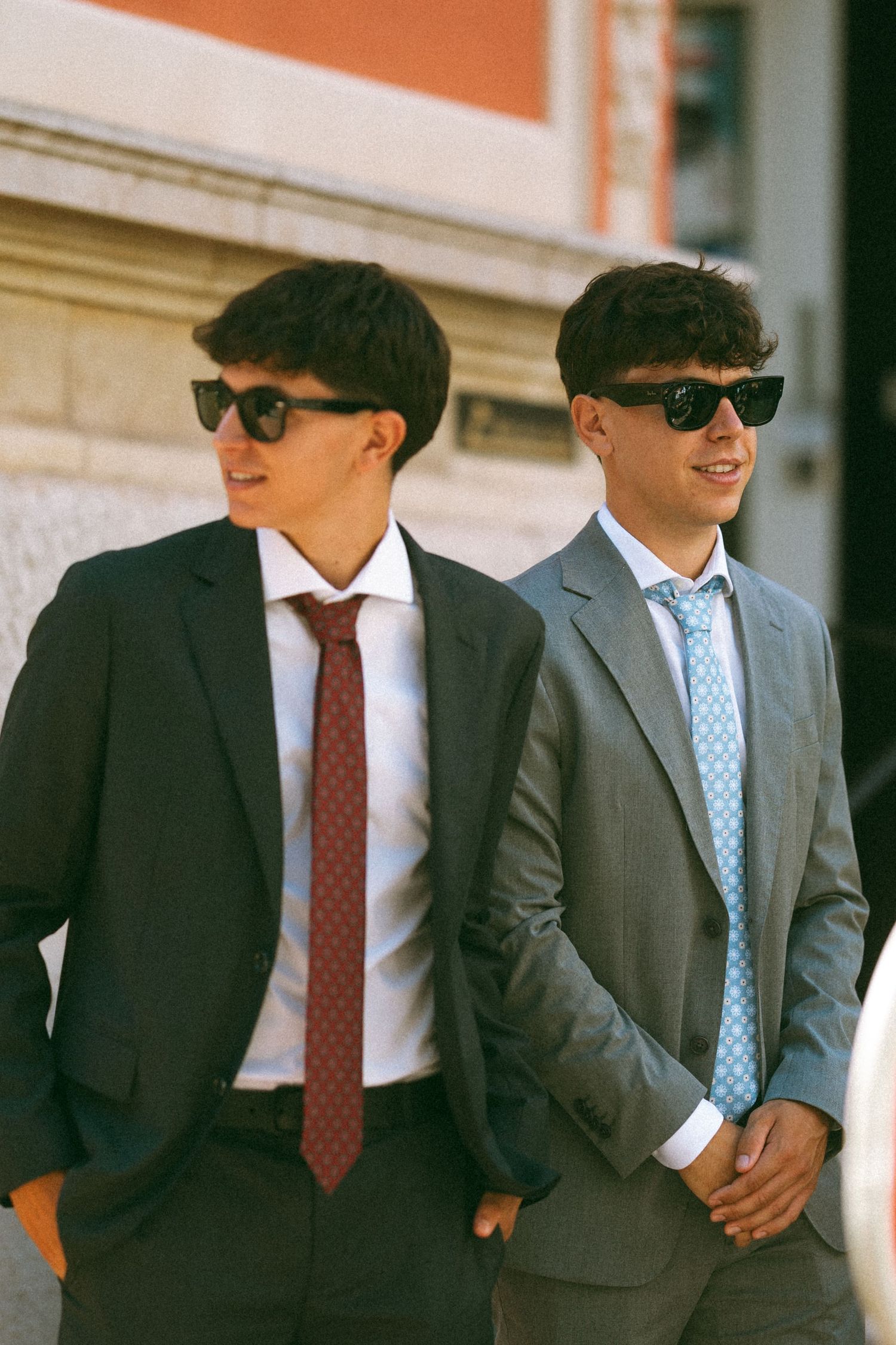 Two young men in suits and sunglasses standing outdoors, one wearing a dark suit with red tie, other in gray suit.