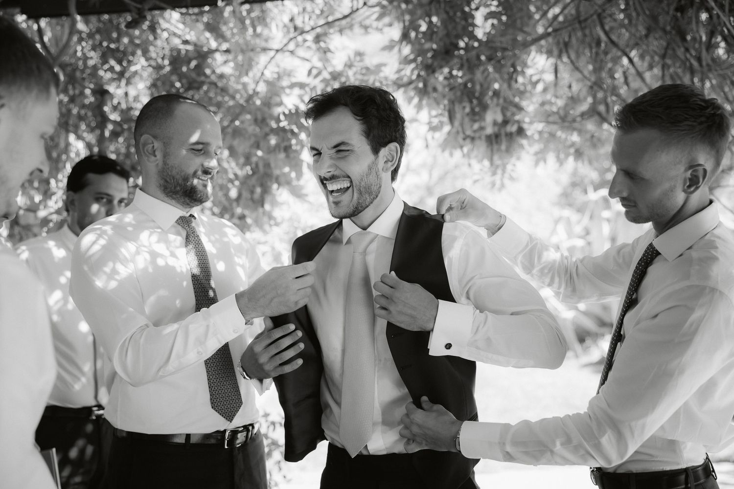 Groomsmen helping groom adjust suit jacket outdoors in black and white wedding photo.