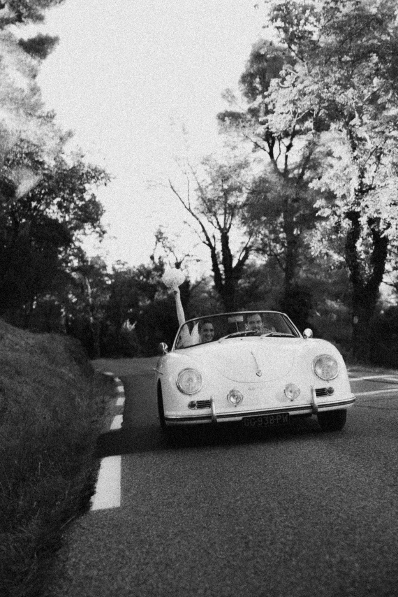 Black and white photo of a vintage white Porsche 356 convertible driving on a tree-lined road with passengers inside.