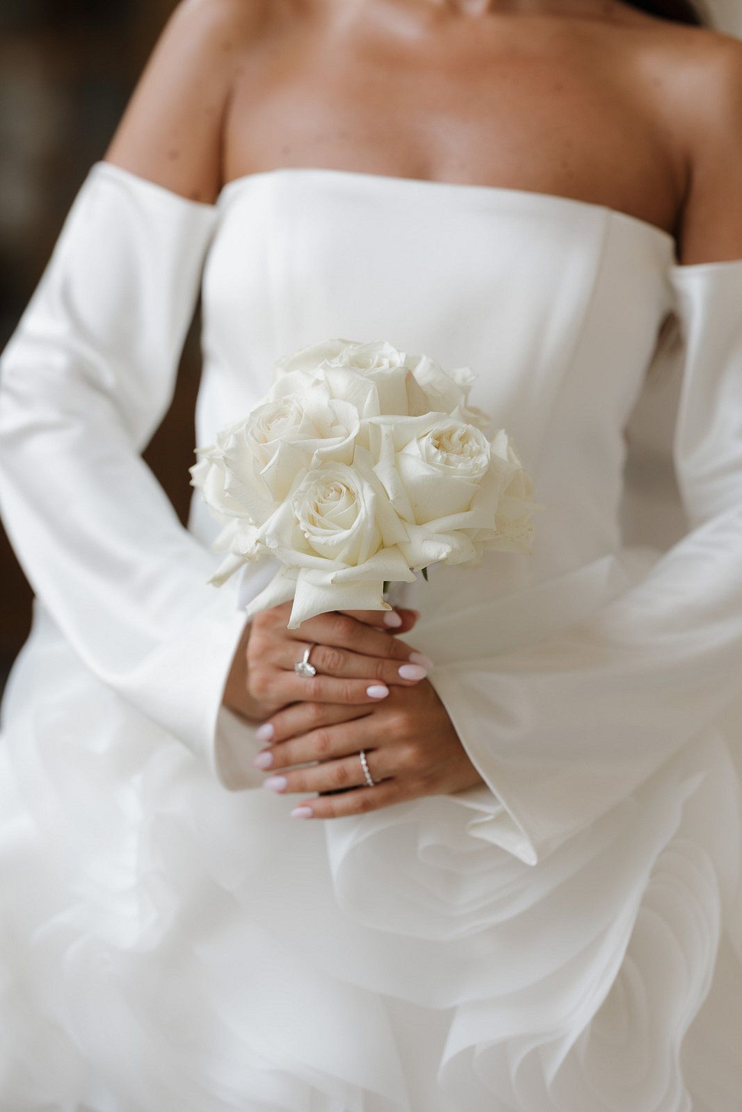 Close-up details of white roses held against an elegant white wedding dress showing bridal hands with rings.