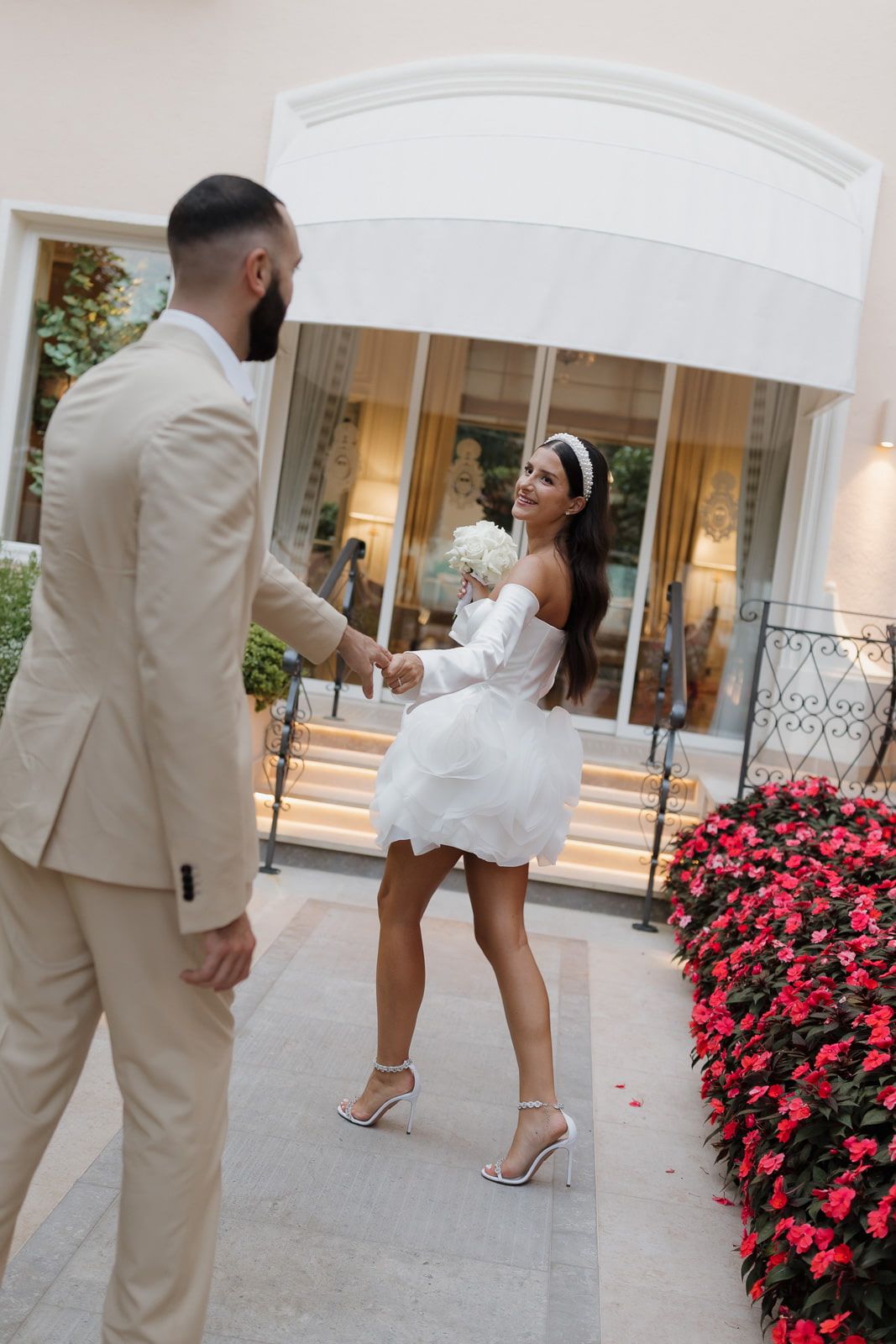Romantic couple in beige and white outfits walking through a courtyard with vibrant pink flowers.