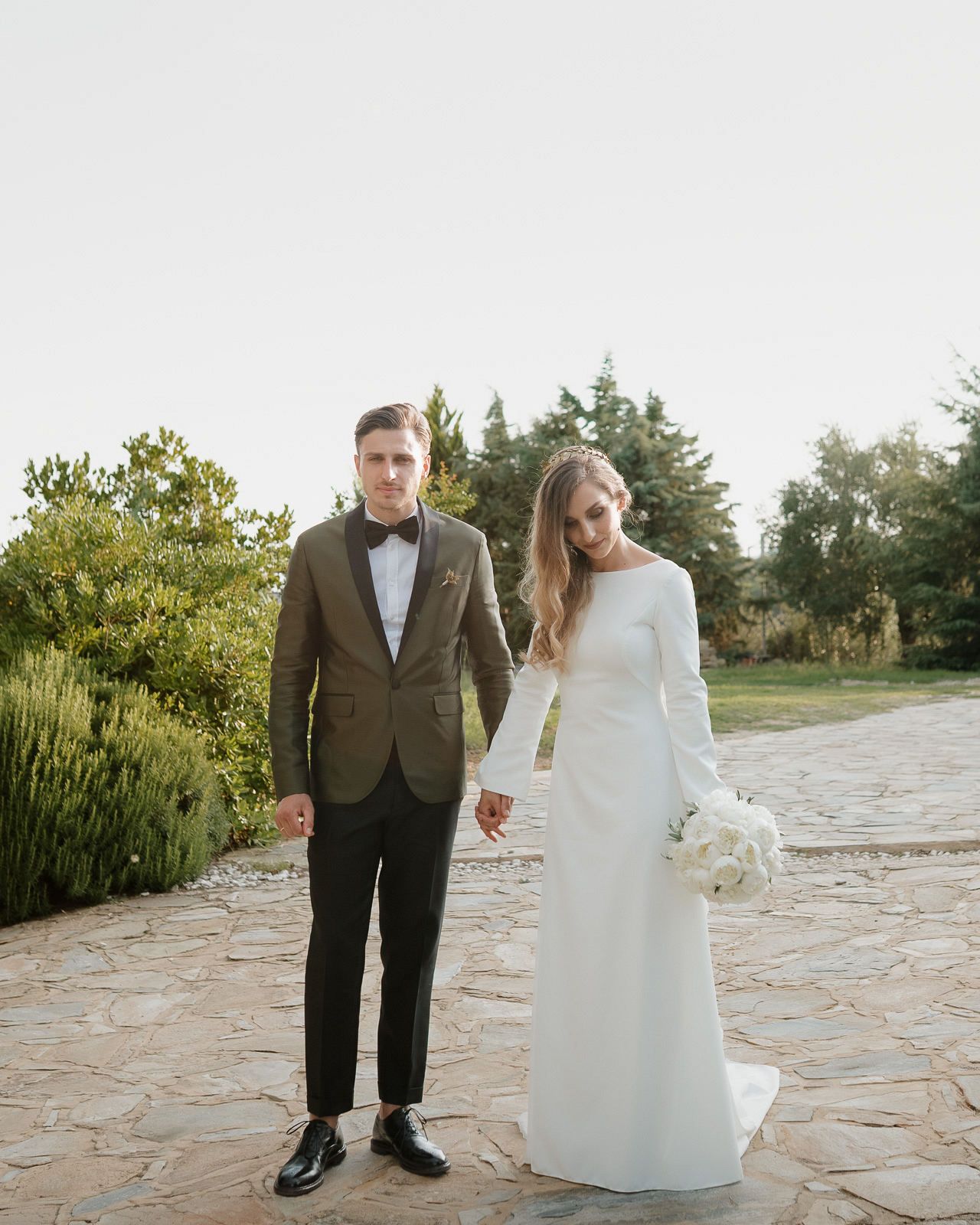 A couple in wedding attire walks hand in hand along a stone pathway surrounded by lush greenery and sunlit trees.