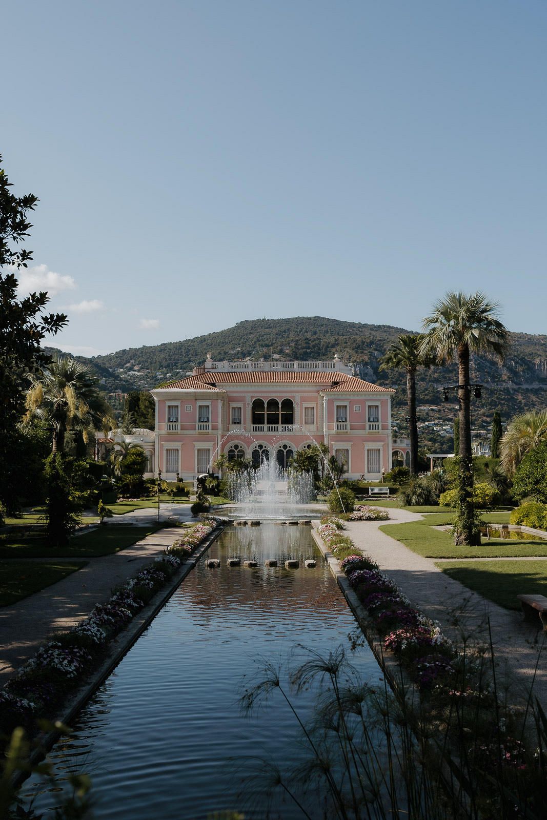 A pink Mediterranean-style villa with a grand fountain pool and palm trees set against a mountainous backdrop.