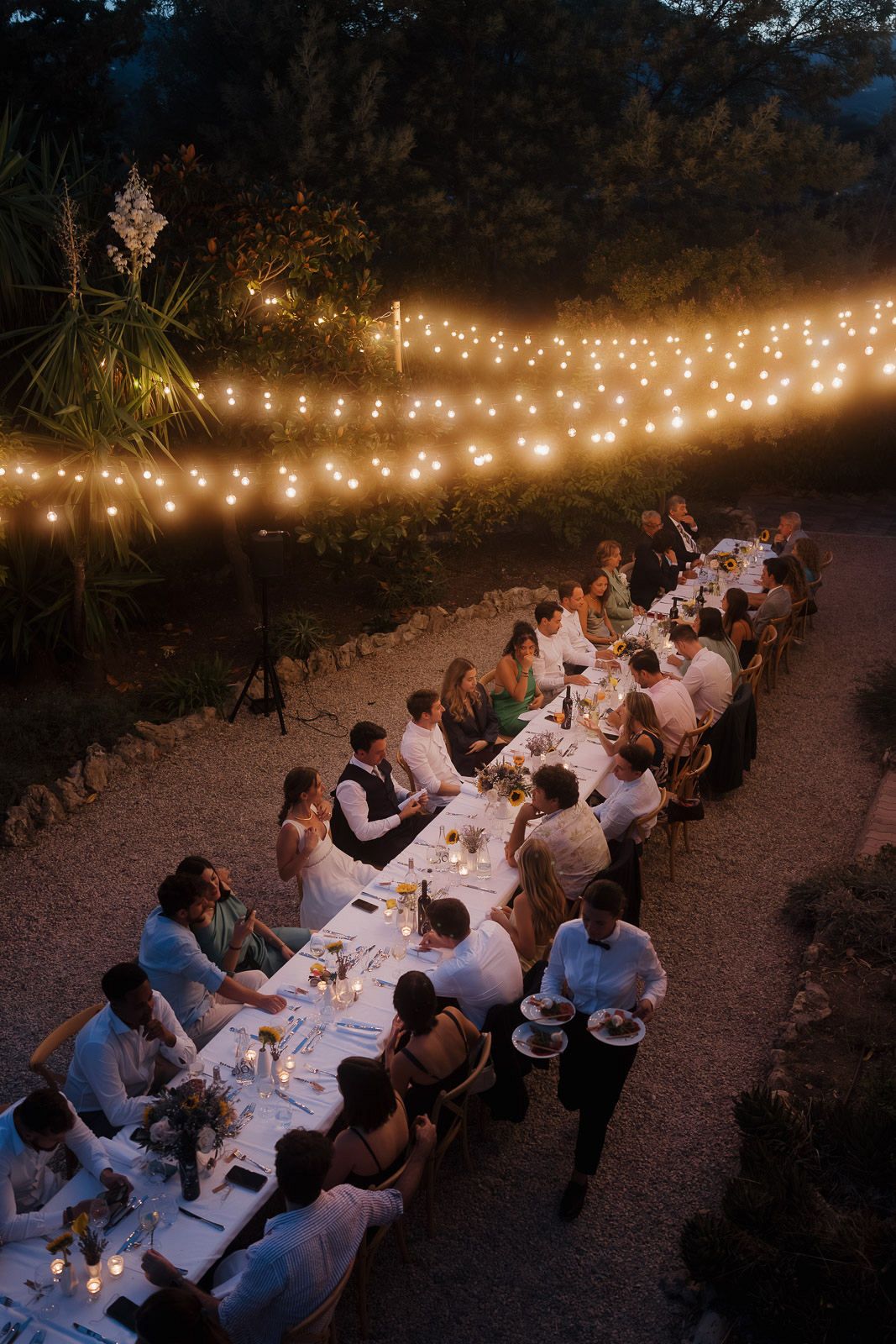 Guests enjoy an intimate outdoor dinner party under warm string lights at dusk with long tables set in nature.