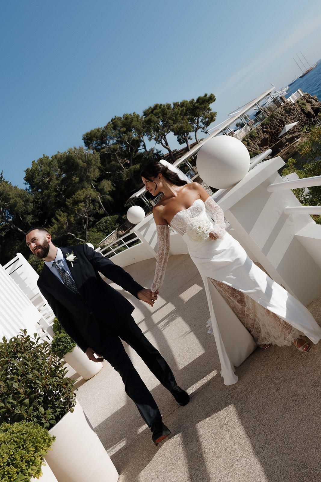 A bride in a white dress and groom in black suit pose together outdoors near white decorative elements.