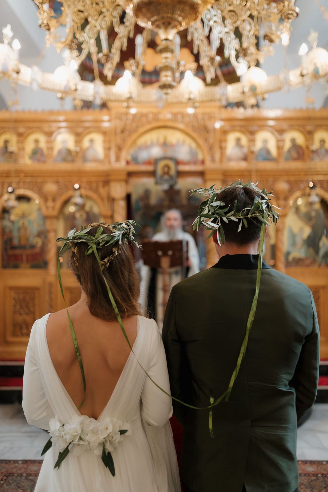 A couple wearing olive leaf crowns stands before an ornate Orthodox church iconostasis during their wedding ceremony.