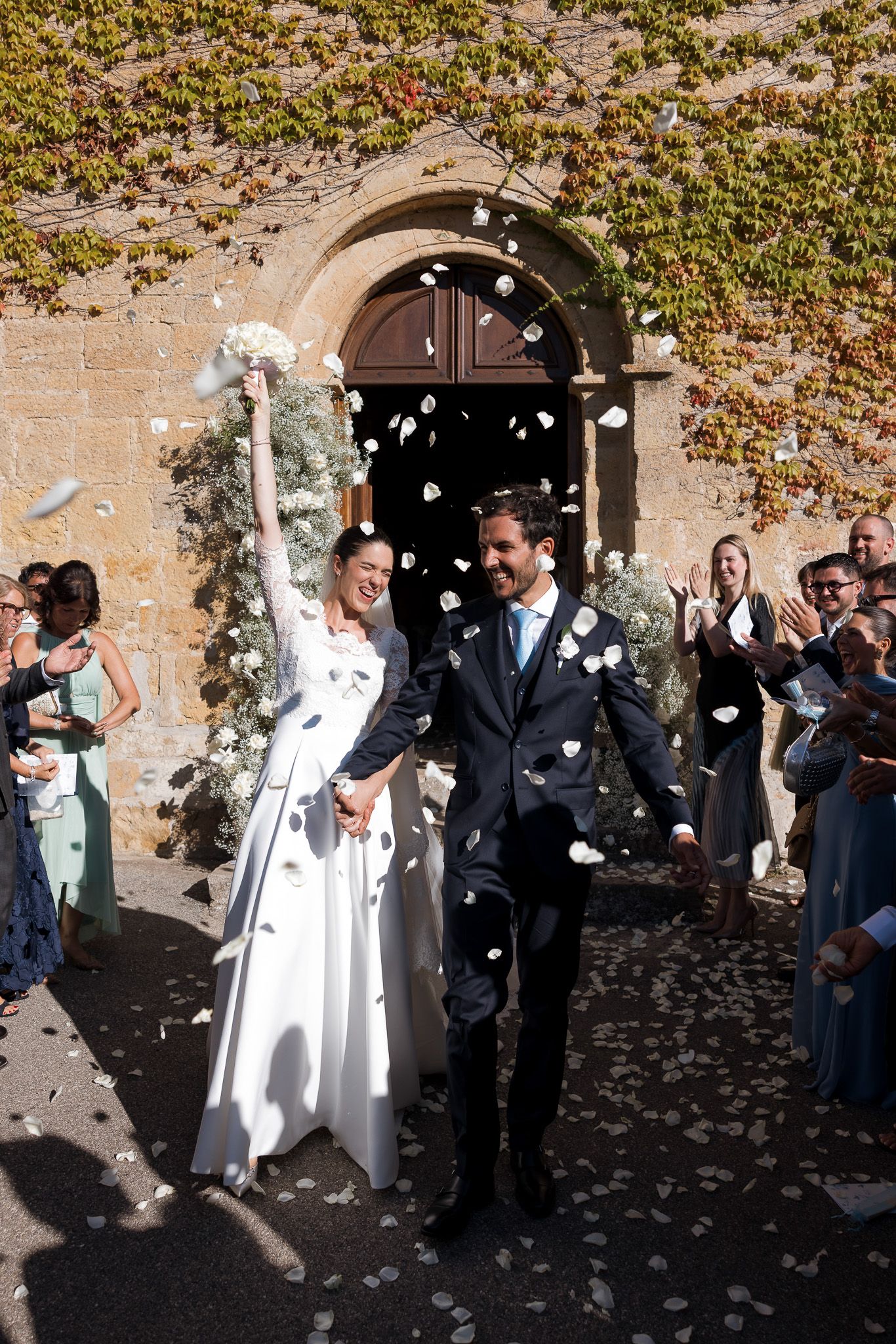 Joyful newlyweds exit church as guests shower them with white flower petals confetti on sunny wedding day.