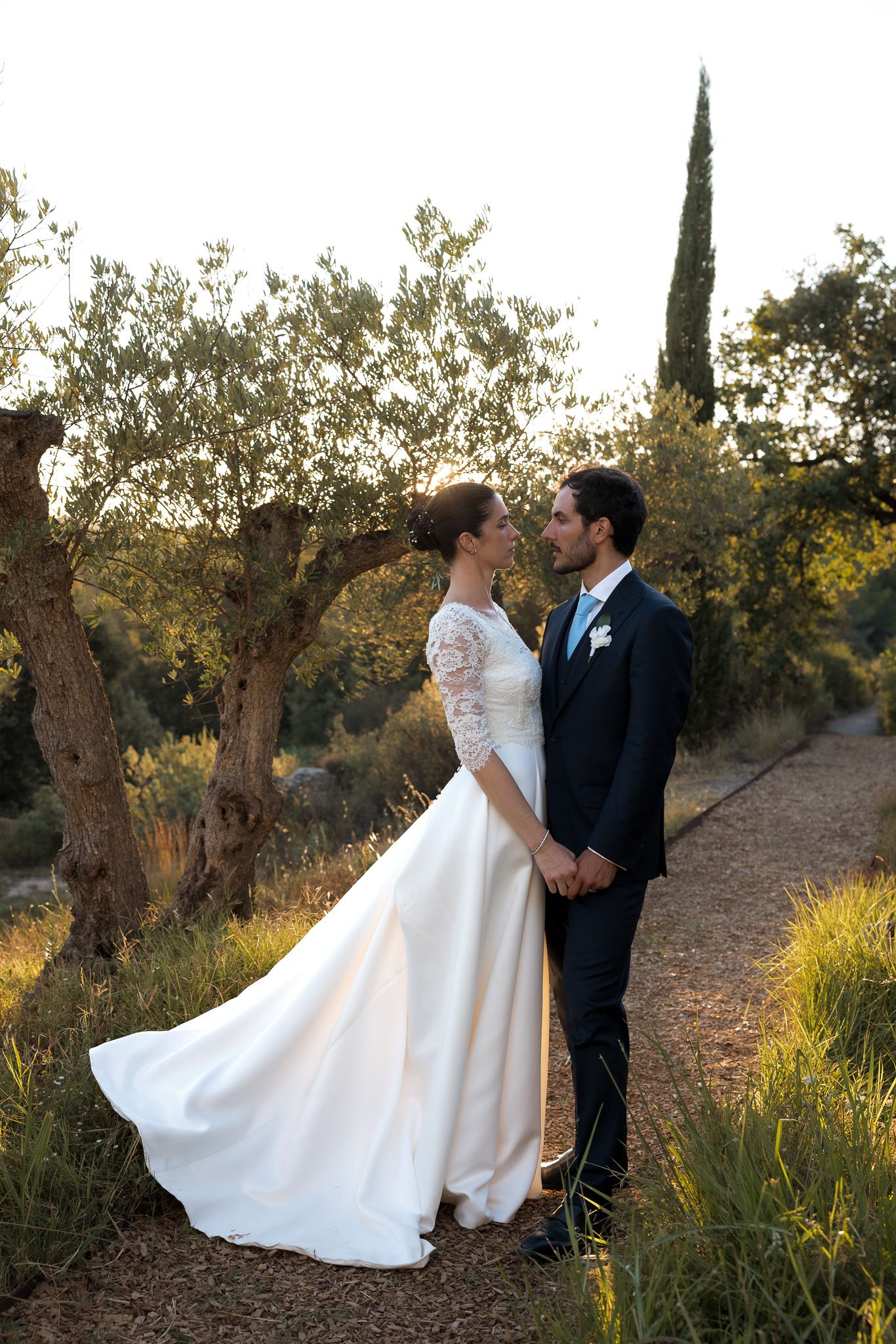 Bride and groom share a romantic moment on a garden path surrounded by olive trees at golden hour.