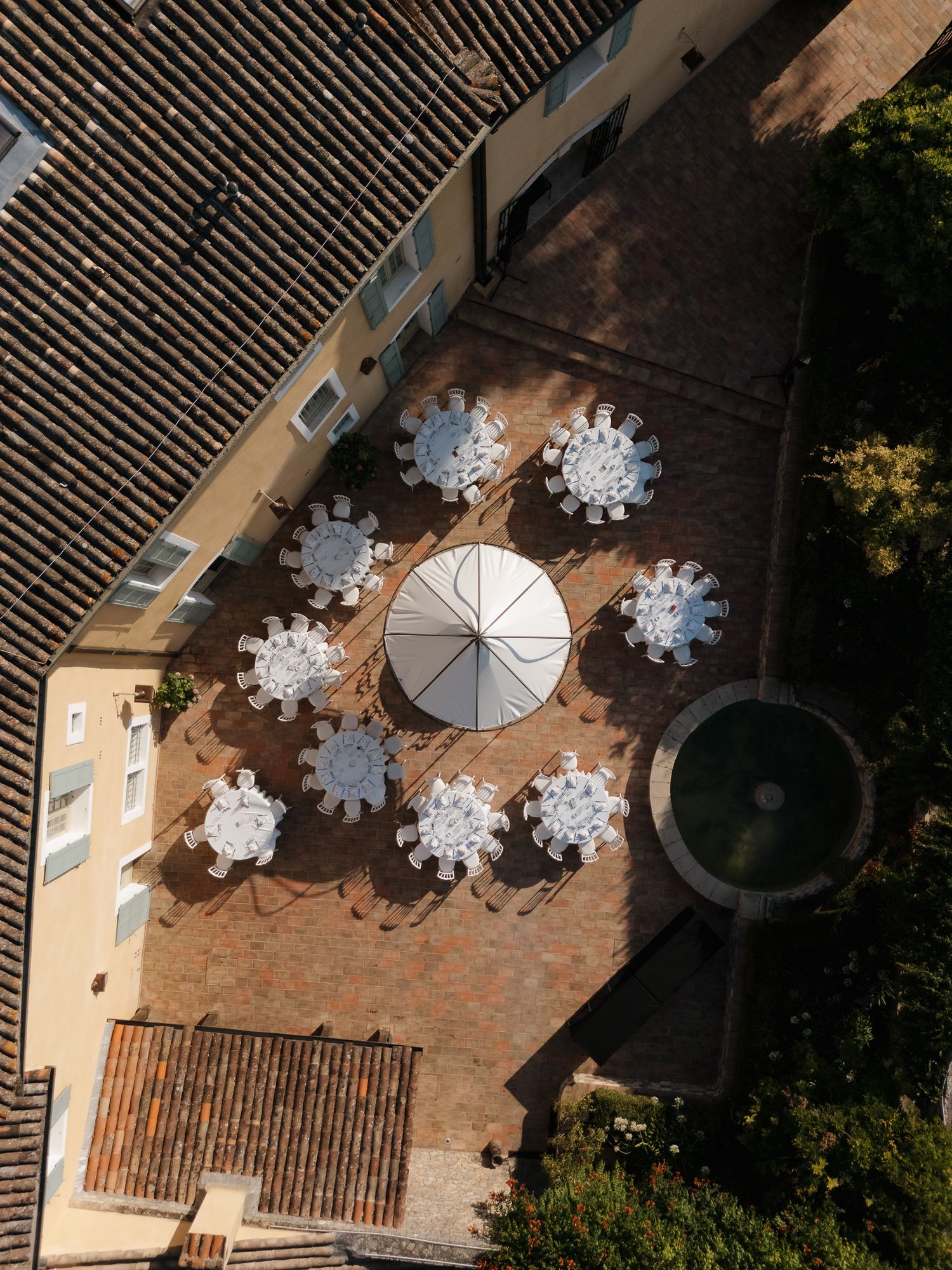 Aerial view of an elegant outdoor courtyard with round white-clothed tables, a large umbrella, and a circular fountain.