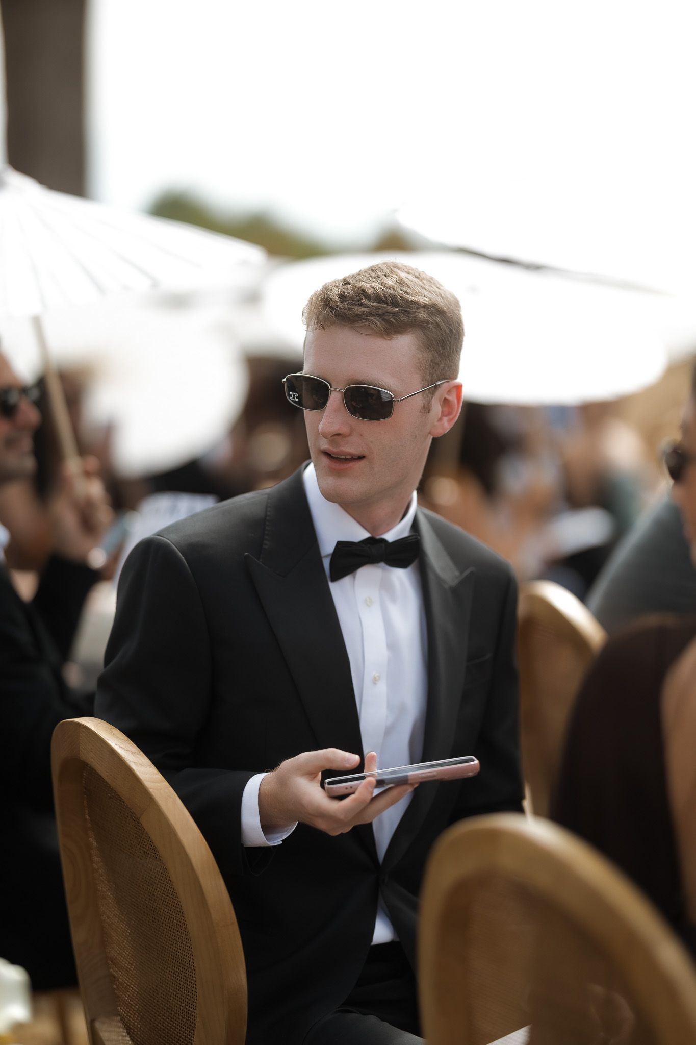 Young man in black tuxedo and sunglasses holding a phone at an outdoor formal event.