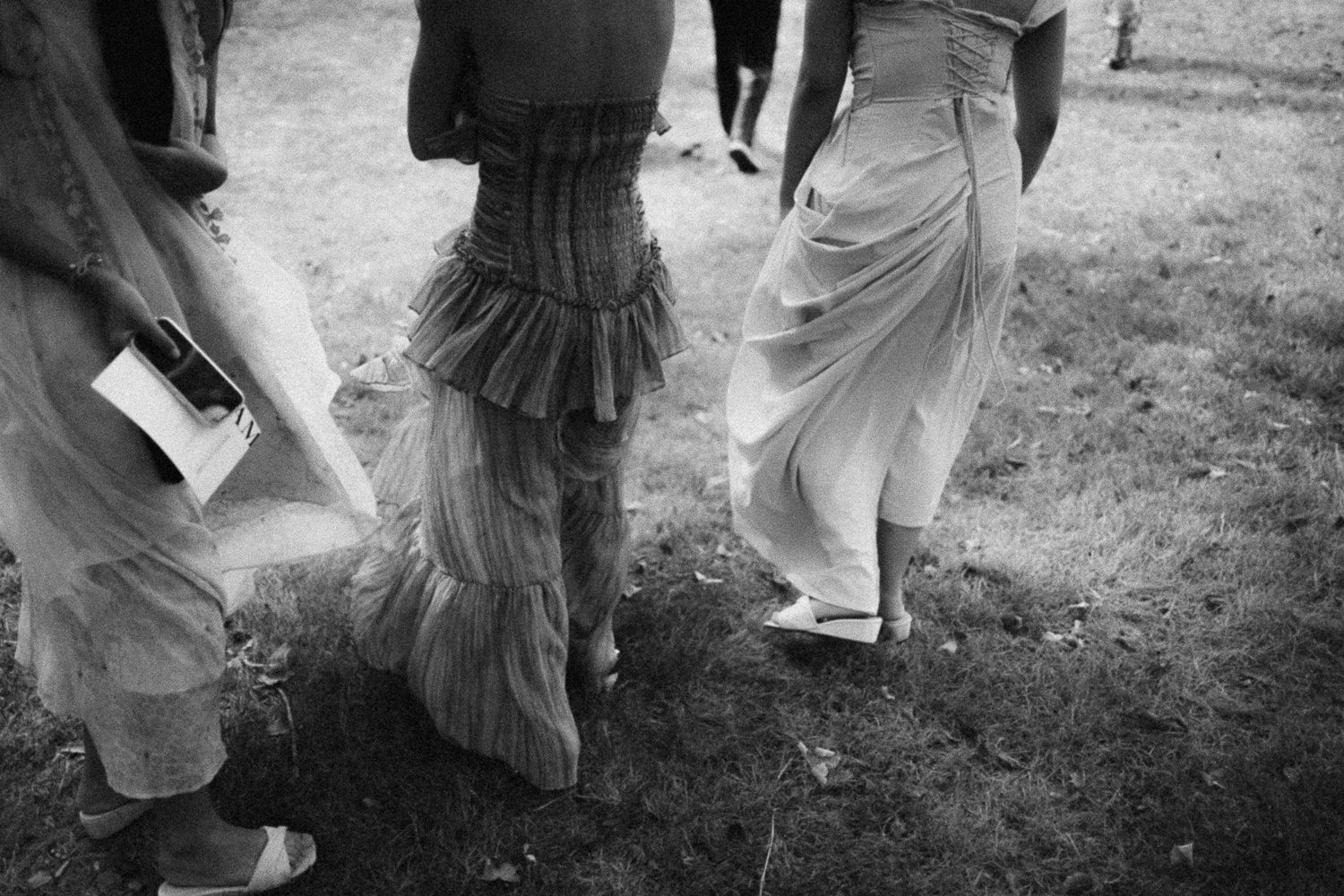 Black and white photo of three women in elegant dresses walking outdoors on grass at a formal event.