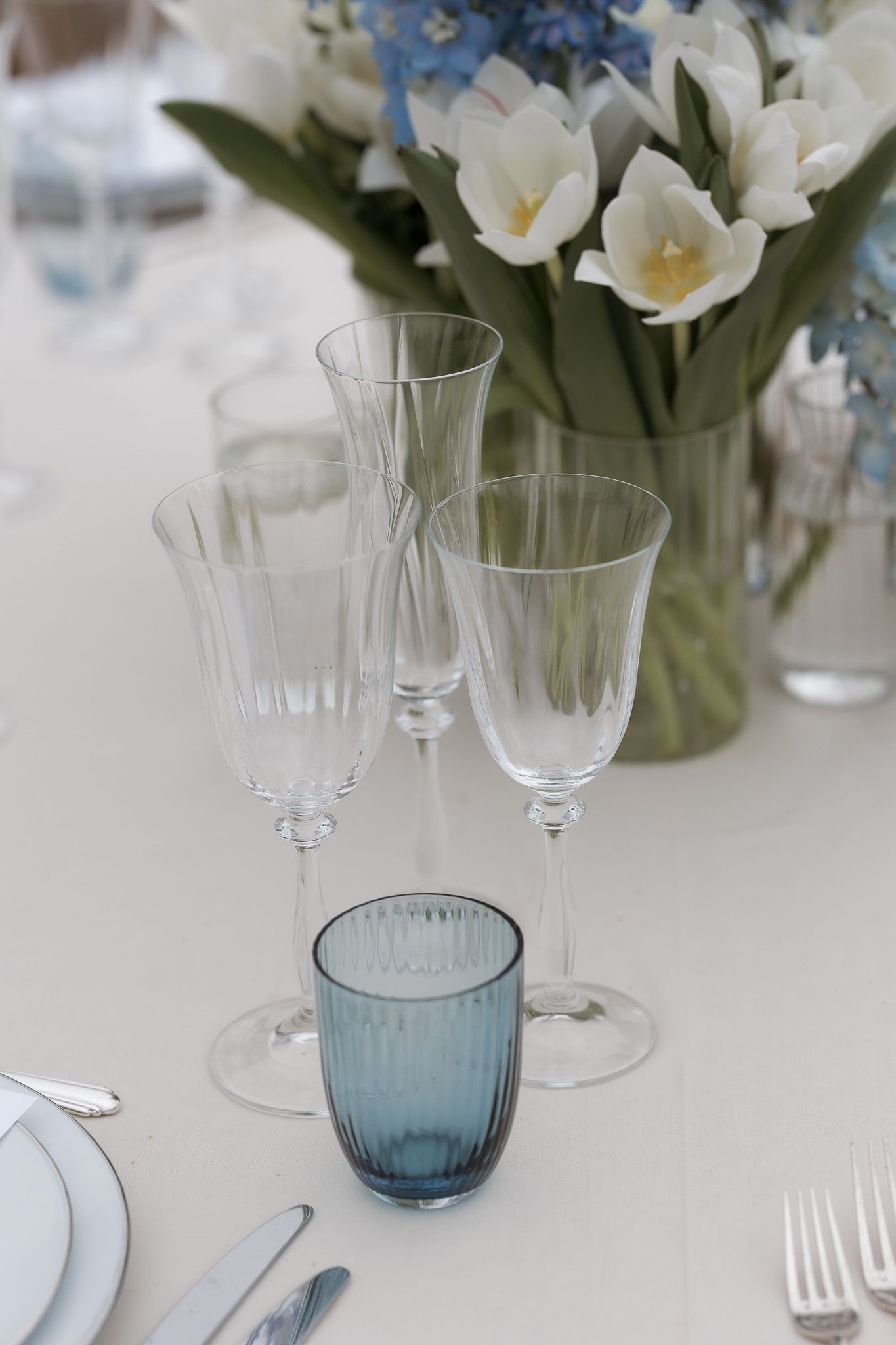 Elegant table setting with crystal wine glasses, blue tumbler, white tulips centerpiece, and silver cutlery on white linen.