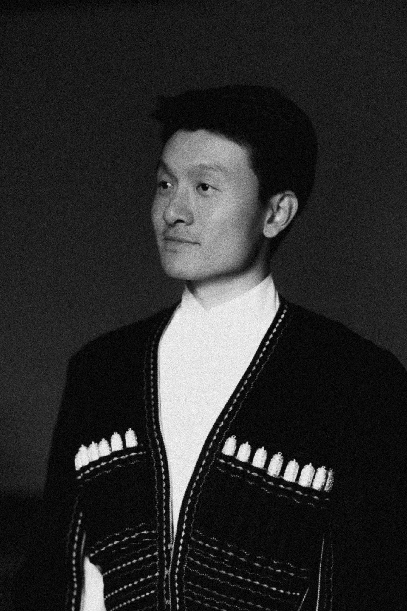 Black and white portrait of a young Asian man wearing a decorative cardigan over a white turtleneck.