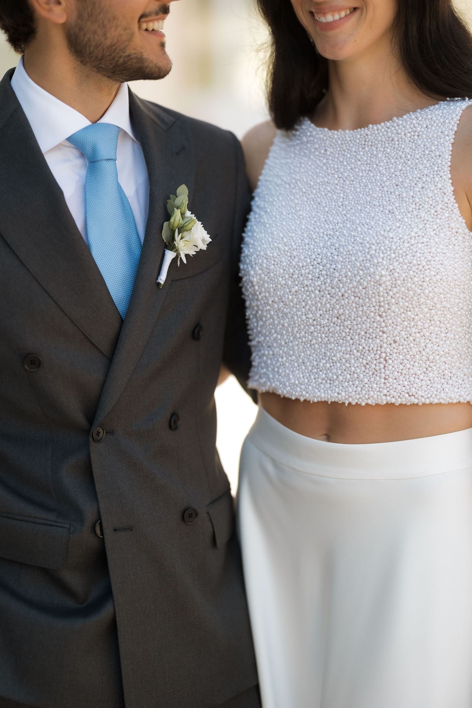 A smiling bride and groom in wedding attire, she wears a pearl crop top and white skirt, he wears a dark suit with blue tie.