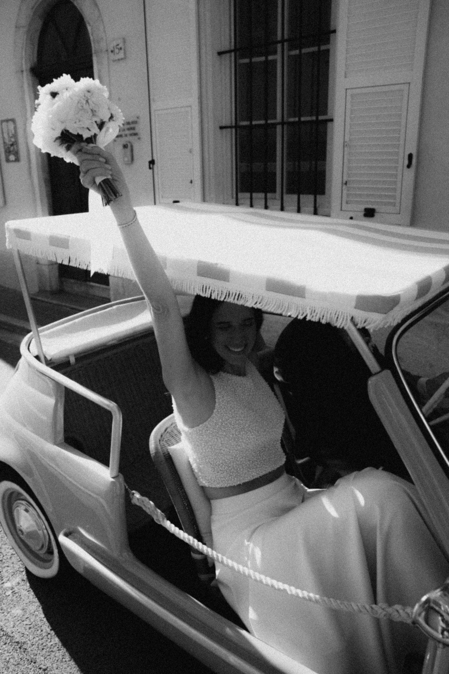 Joyful bride in white dress raising bouquet from vintage convertible car, black and white wedding photo.