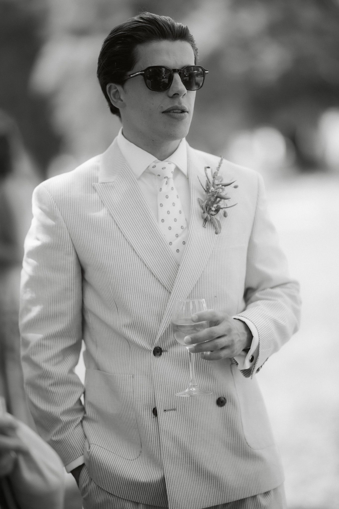 Elegant man in double-breasted suit with boutonniere, sunglasses, holding champagne glass at formal event.