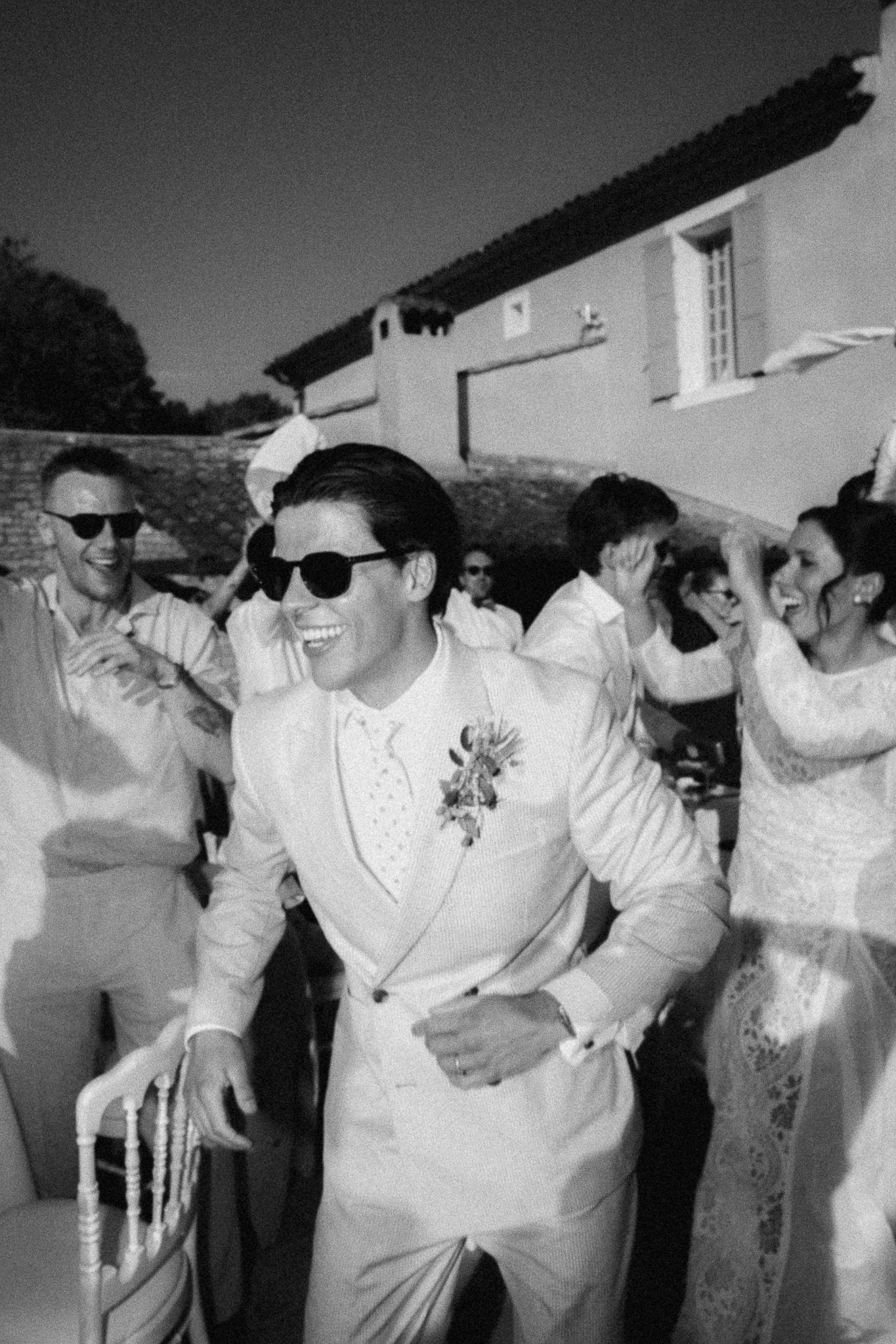 Groom in white suit and sunglasses dancing joyfully with wedding guests outdoors in black and white photo.