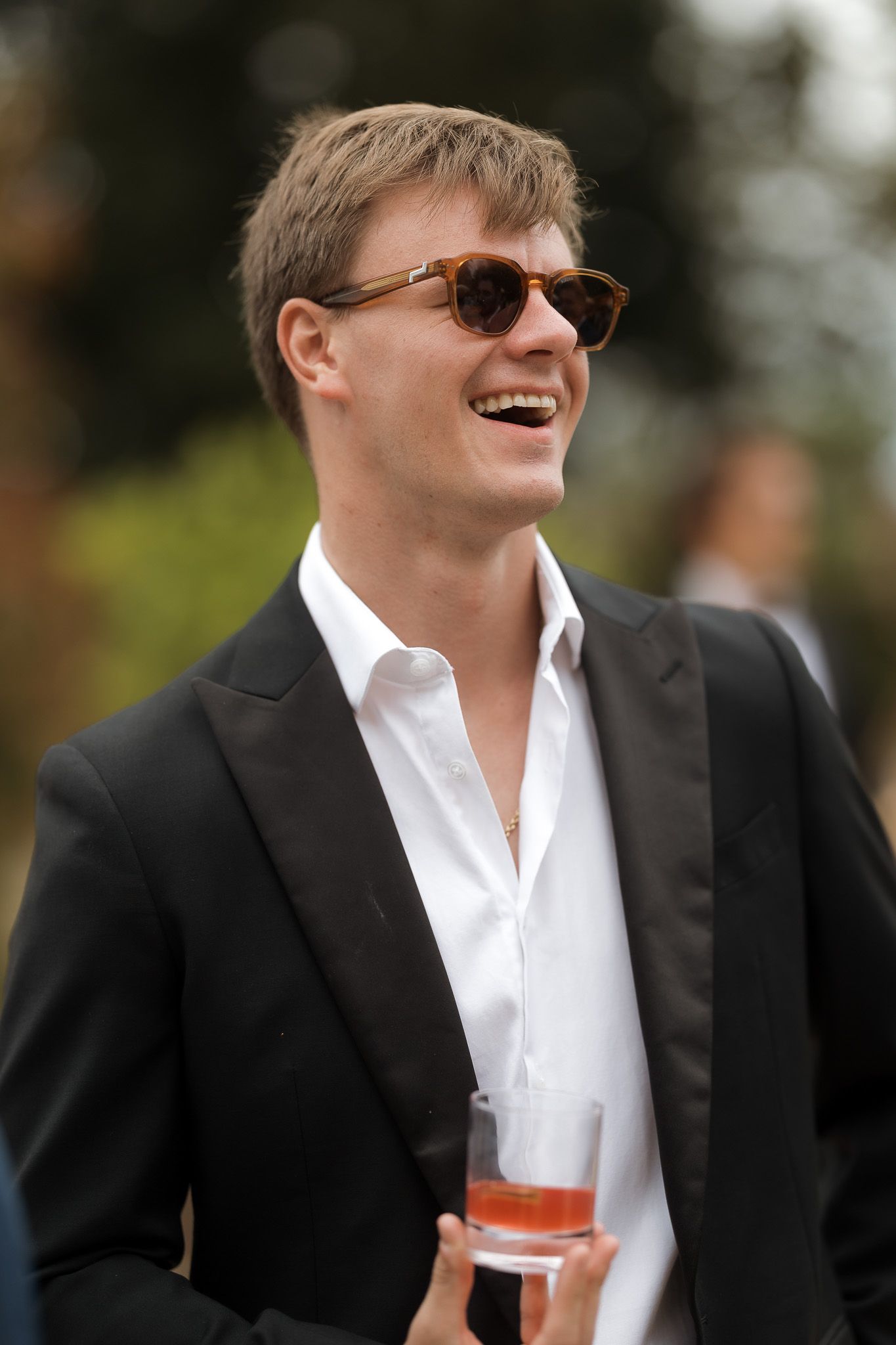 Smiling young man in black suit and sunglasses holding a cocktail at an outdoor event.