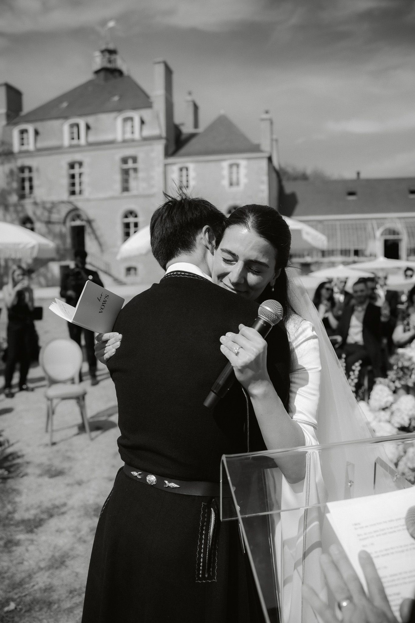 Black and white photo of bride and groom sharing an emotional hug during outdoor wedding ceremony at elegant French château.